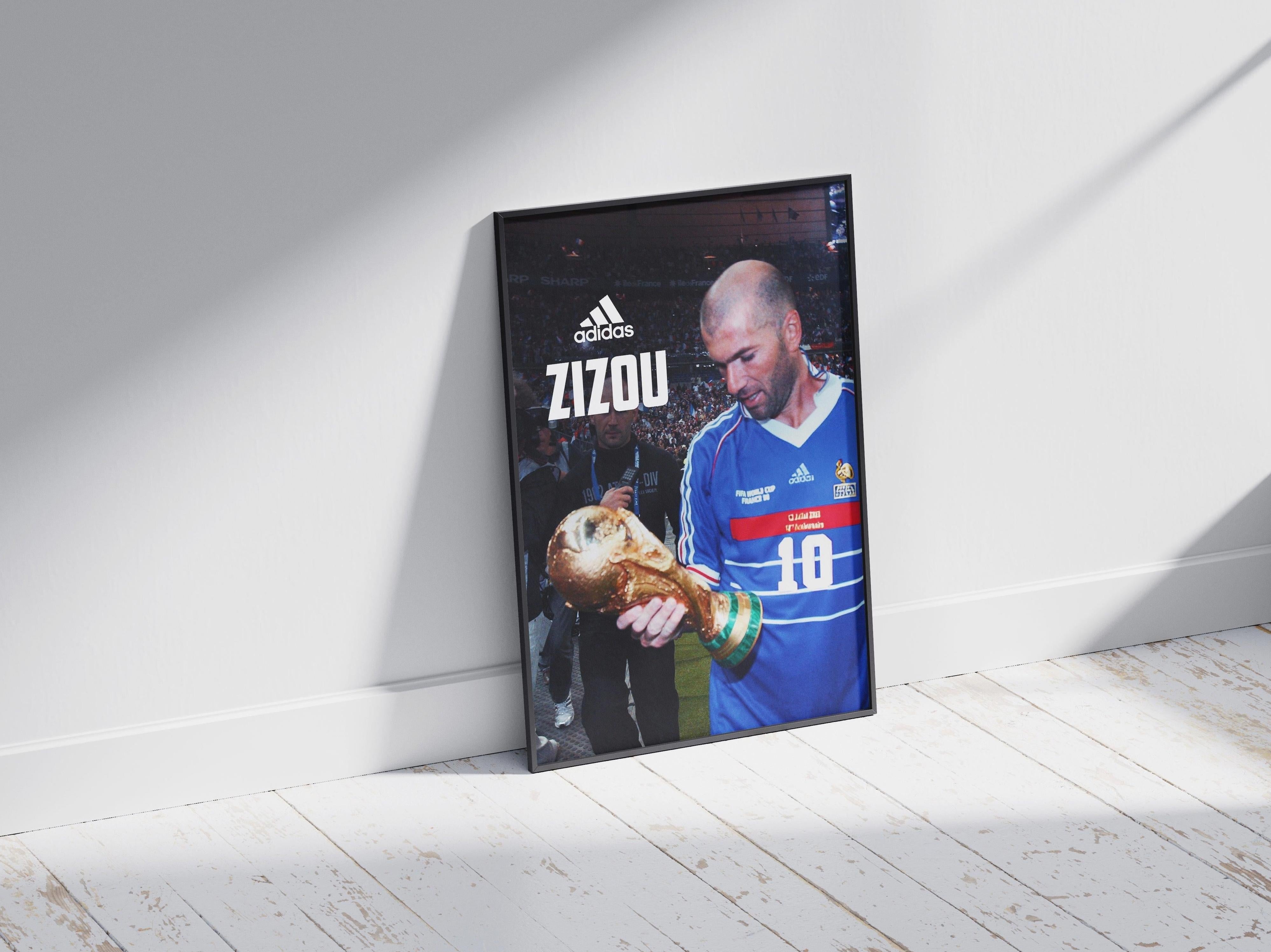 The Retro1999 Zinedine Zidane Zizou Poster features Zidane in a blue France jersey holding the World Cup, with ADIDAS and ZIZOU above. It leans against a white wall on a wooden floor, sunlight streaming in.