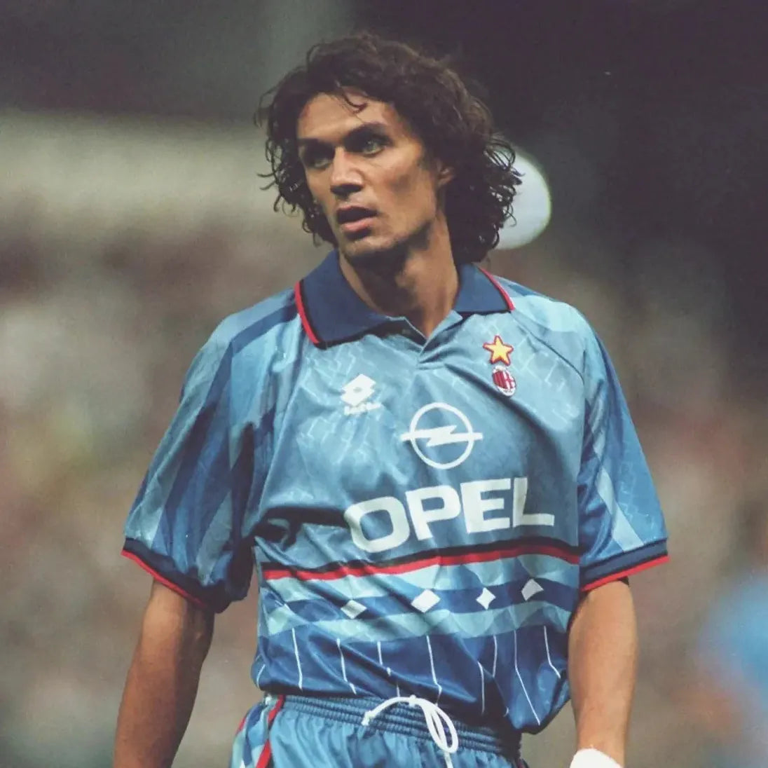 A male soccer player with long curly hair wears the Retro1999 AC Milan 1995-96 Third Kit in light blue with the Opel logo, standing on the field and showcasing this authentic retro design.