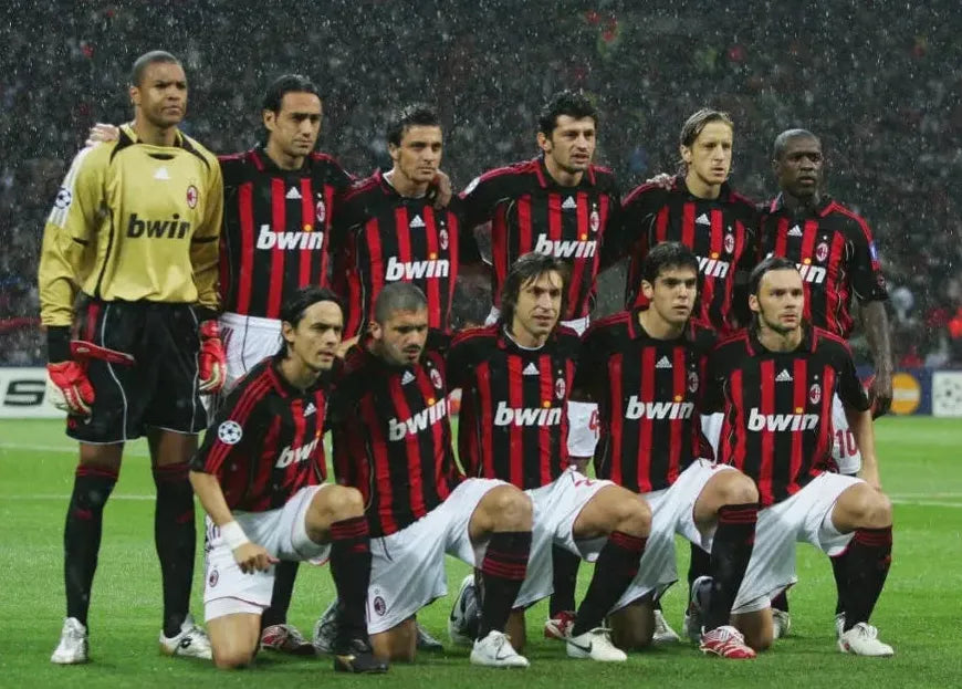 Eleven AC Milan players in classic red and black stripes pose for a team photo on a rainy field, proudly wearing the Retro1999 AC Milan 2006-07 Home Shirt.