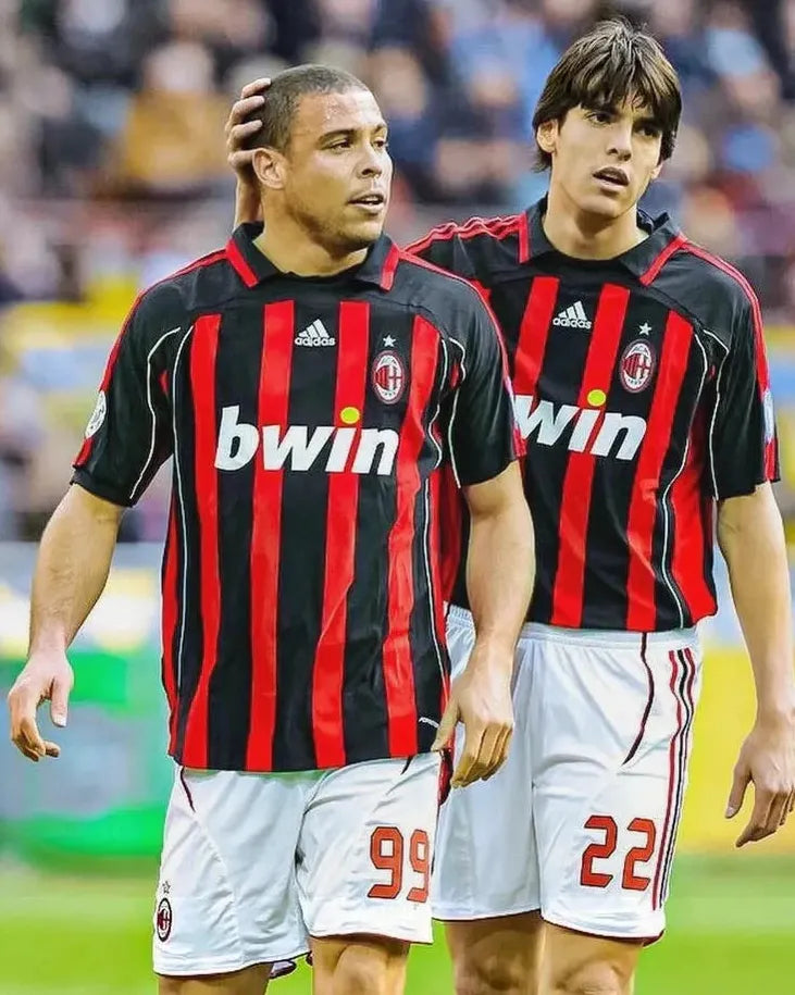 Two soccer players wear the Retro1999 AC Milan 2006-07 Home Shirt, standing side by side on the field—one with a hand on the others head—as they focus during an intense match in classic red and black stripes.