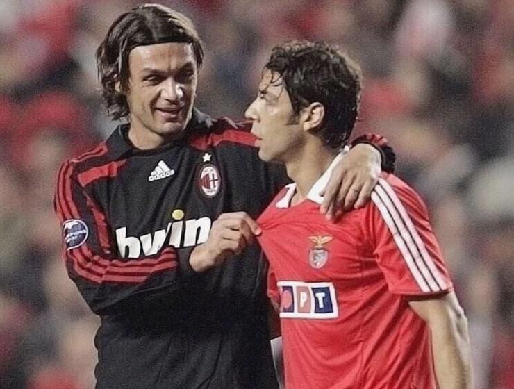 A soccer player in a black Retro1999 AC Milan 2007-08 Third Shirt smiles and puts his arm around a player in a red Benfica jersey as they chat on the field, celebrating football history.
