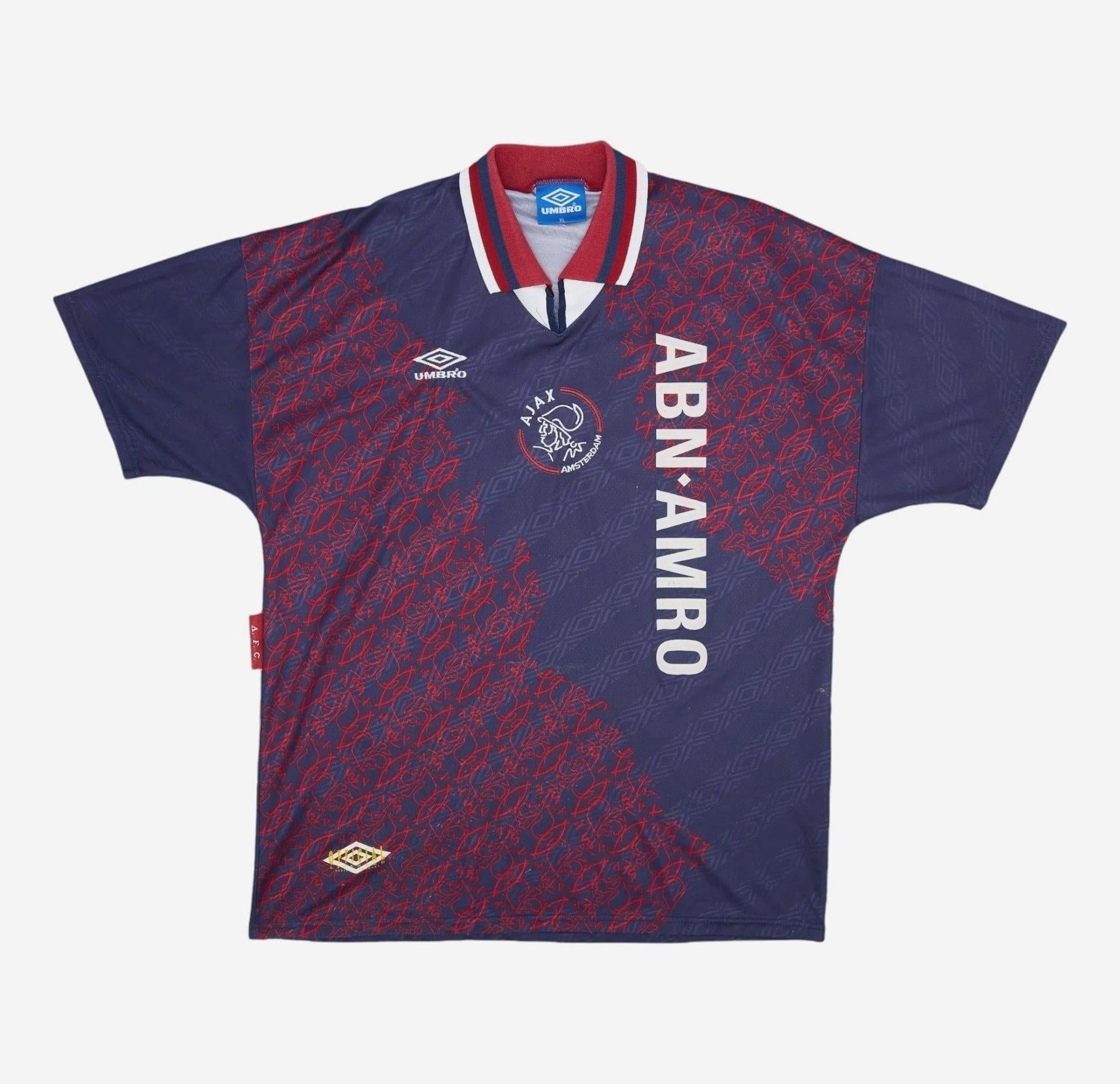 The Retro1999 Ajax 1994-95 Away Shirt features a vintage dark blue and red pattern, white “ABN AMRO” sponsor, Ajax crest, Umbro logo on the chest, and red, white, and blue collar trim—a classic for football fans.