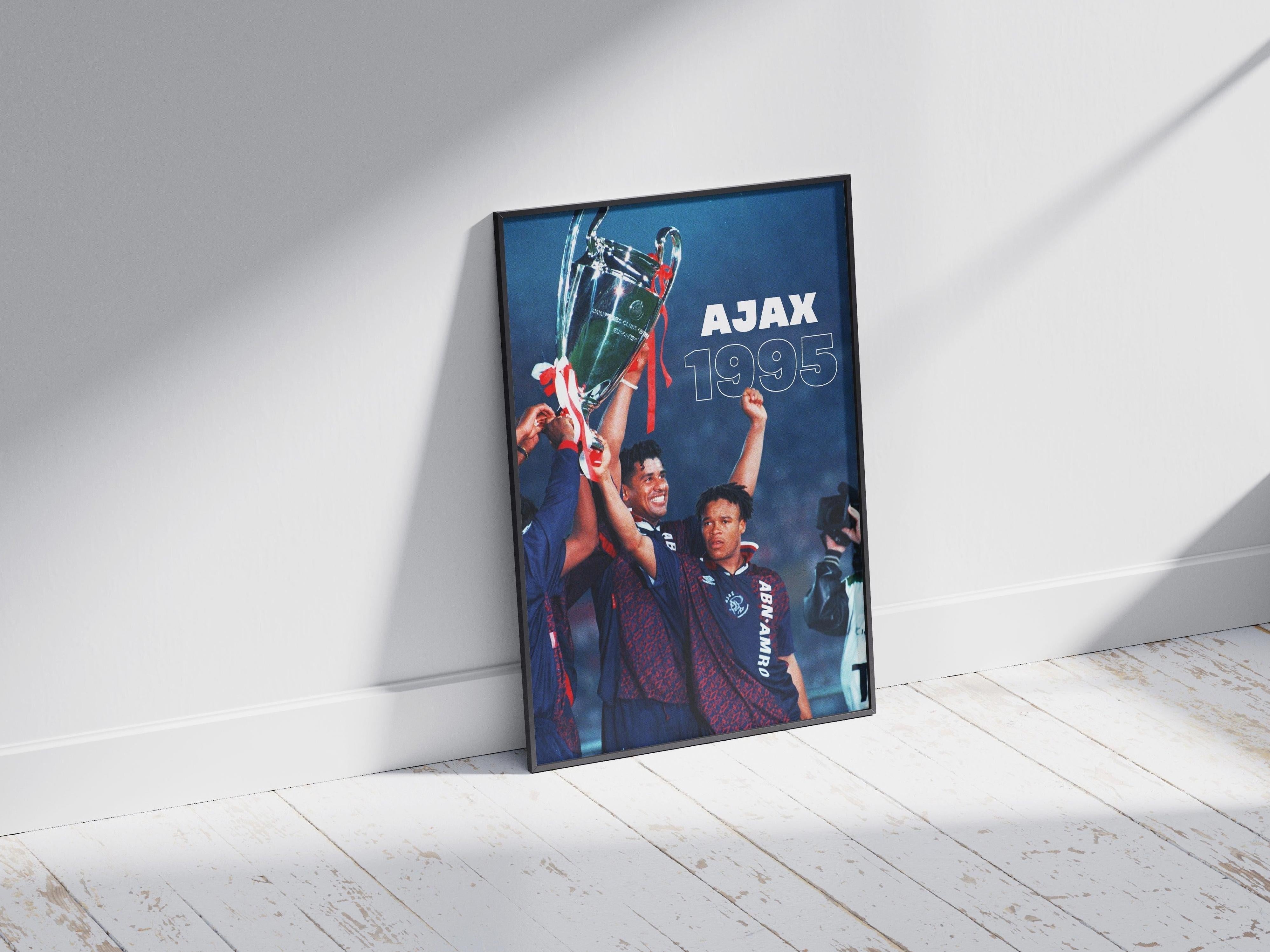 The Retro1999 Ajax 1995 Champions League Poster shows three players celebrating their iconic trophy win, framed and resting on a wooden floor with sunlight casting striking shadows—an ideal piece of football memorabilia.
