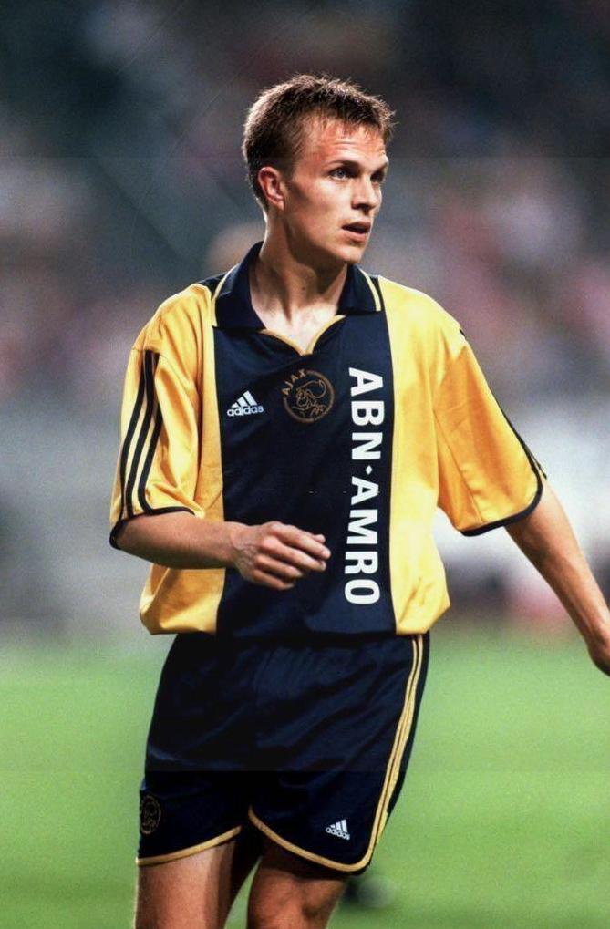 A male soccer player wears the Retro1999 Ajax 2000-01 Away Shirt with ABN AMRO sponsor on the field during a match, gazing left. The background is blurred, with Ajax supporters visible in the distance.