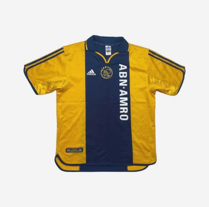 The Ajax 2000-01 Away Shirt by Retro1999 is a yellow and navy retro kit with ABN-AMRO sponsor, Ajax Amsterdam logo, Adidas branding, and signature three shoulder stripes—a fan favorite.