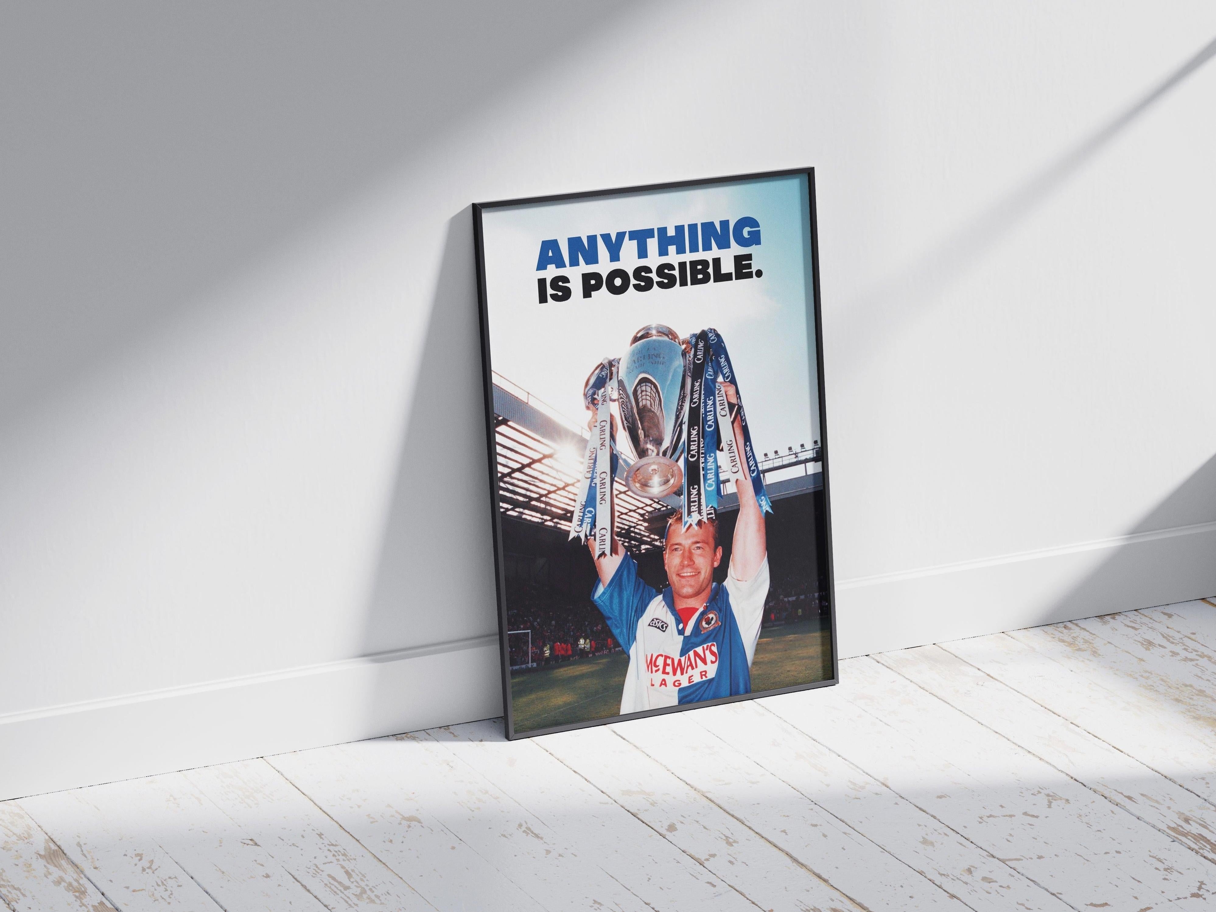 The Retro1999 Alan Shearer Anything Is Possible Poster shows the Blackburn Rovers legend in blue and white holding a trophy, with bold text above. Framed, it rests on a white wooden floor as sunlight casts shadows across this iconic football moment.