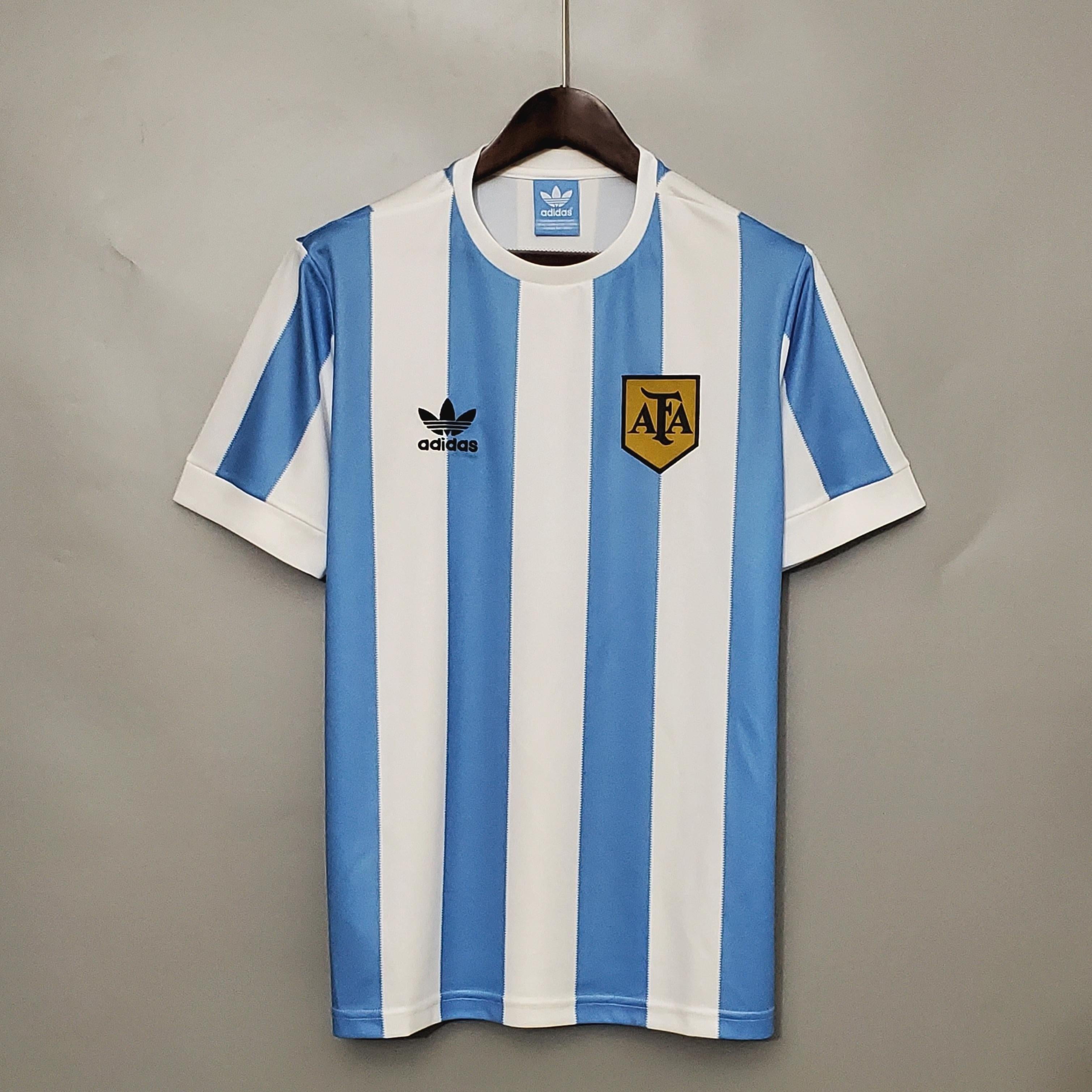 A blue and white vertically striped Argentina 1978 Home Shirt by Retro1999 hangs on a hanger against a gray wall, featuring classic retro details inspired by the iconic football kit.