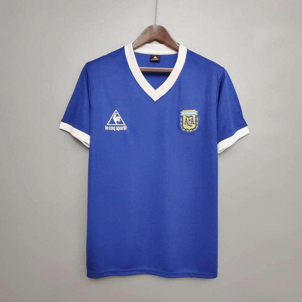 The Retro1999 Argentina 1986 Away Shirt is a blue soccer jersey with white collar and sleeve edges, featuring the AFA crest, hanging on a wooden hanger against a light background.