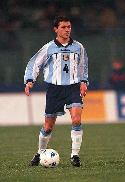An athlete in a Retro1999 Argentina 2000/01 Home Shirt, number 4, controls the ball on a grass pitch during a match. Stadium lights glow in the blurred background.