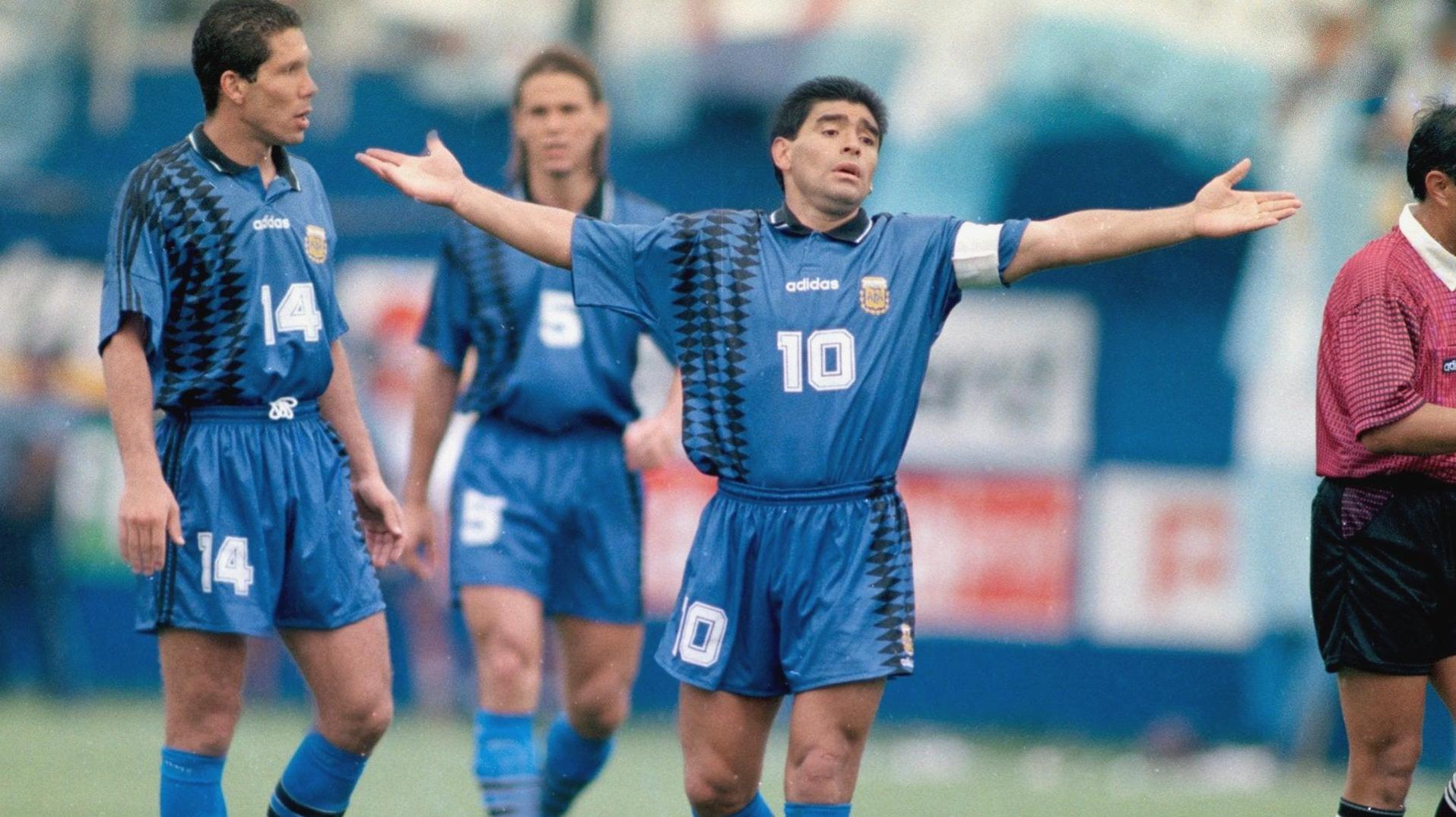 Three soccer players wear the Retro1999 Argentina Retro 1994 Away Shirt on the field, with number 10 in the center, arms outstretched and looking frustrated—a memorable World Cup 1994 moment. A referee in pink is partly visible.