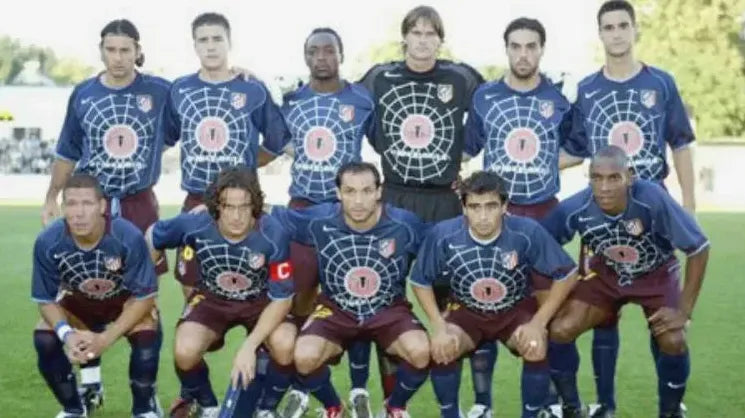 Eleven soccer players pose on a field, wearing the Retro1999 Atletico Madrid 2004-05 Away Shirt with a bold spiderweb and target design, arranged in two rows with some kneeling in front for a team photo.