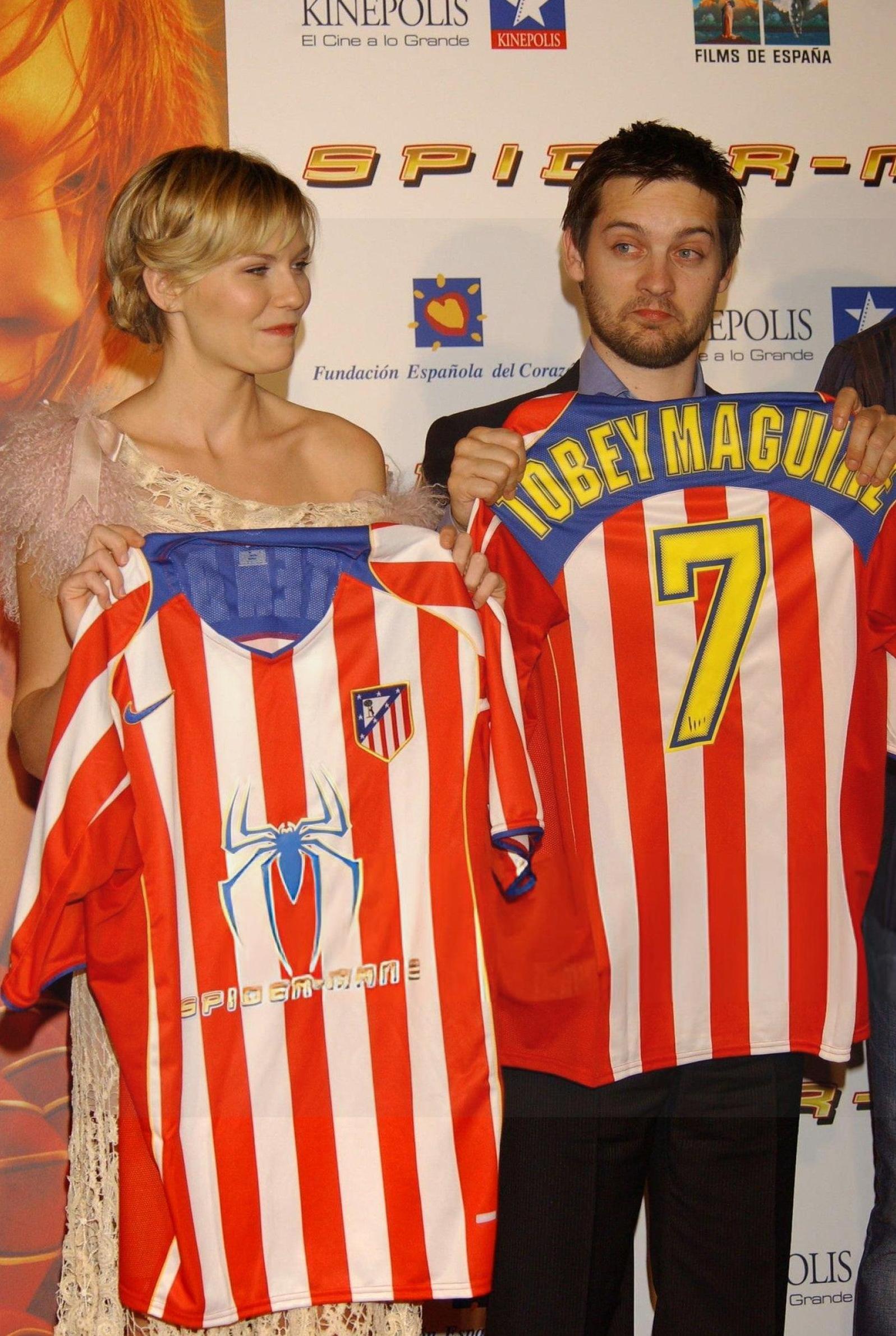 At an event, two people hold Retro1999 Atletico Madrid 2004-05 Home Shirts featuring Spider-Man logos; one displays Tobey Maguire 7. A Spider-Man movie poster and Columbia Pictures sponsorship signs are seen in the background.