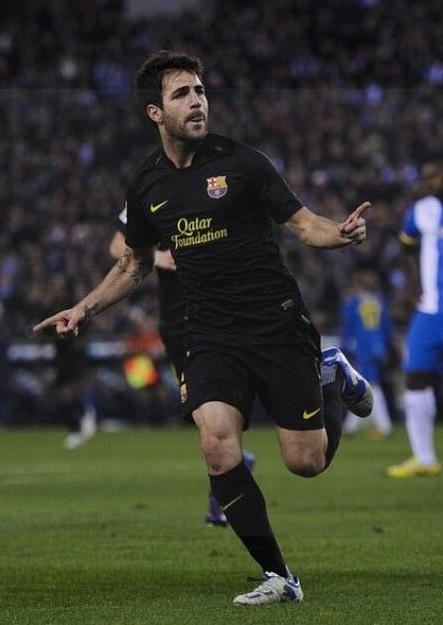 Wearing the Retro1999 Barcelona 2011-12 Away Shirt, a soccer player celebrates on the field with arms outstretched. Other players and a blurred stadium crowd appear in the background.