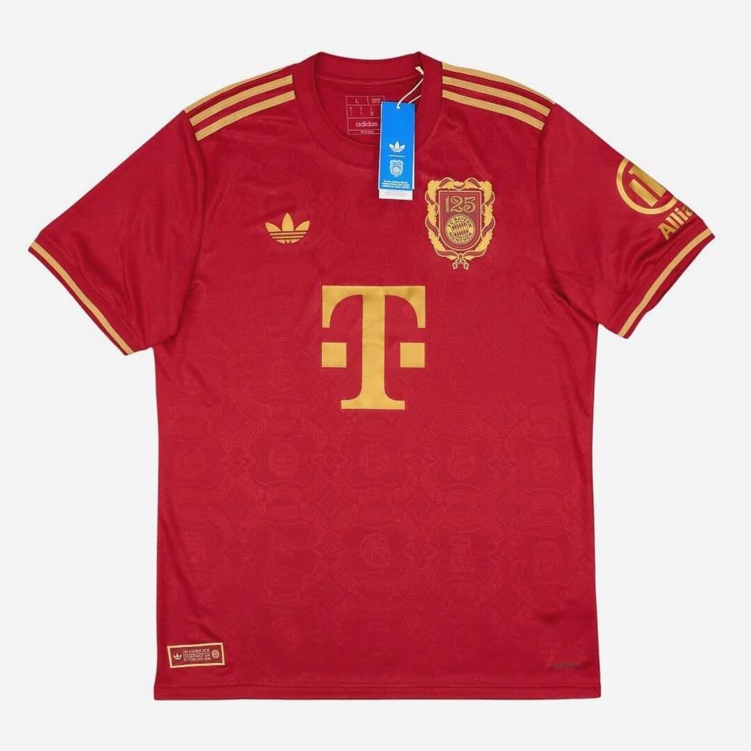 The Retro1999 Bayern Munich 2024-25 125th Anniversary Shirt is red with gold accents, a large T logo, special 125th anniversary crest, gold shoulder stripes, and has tags attached at the collar.