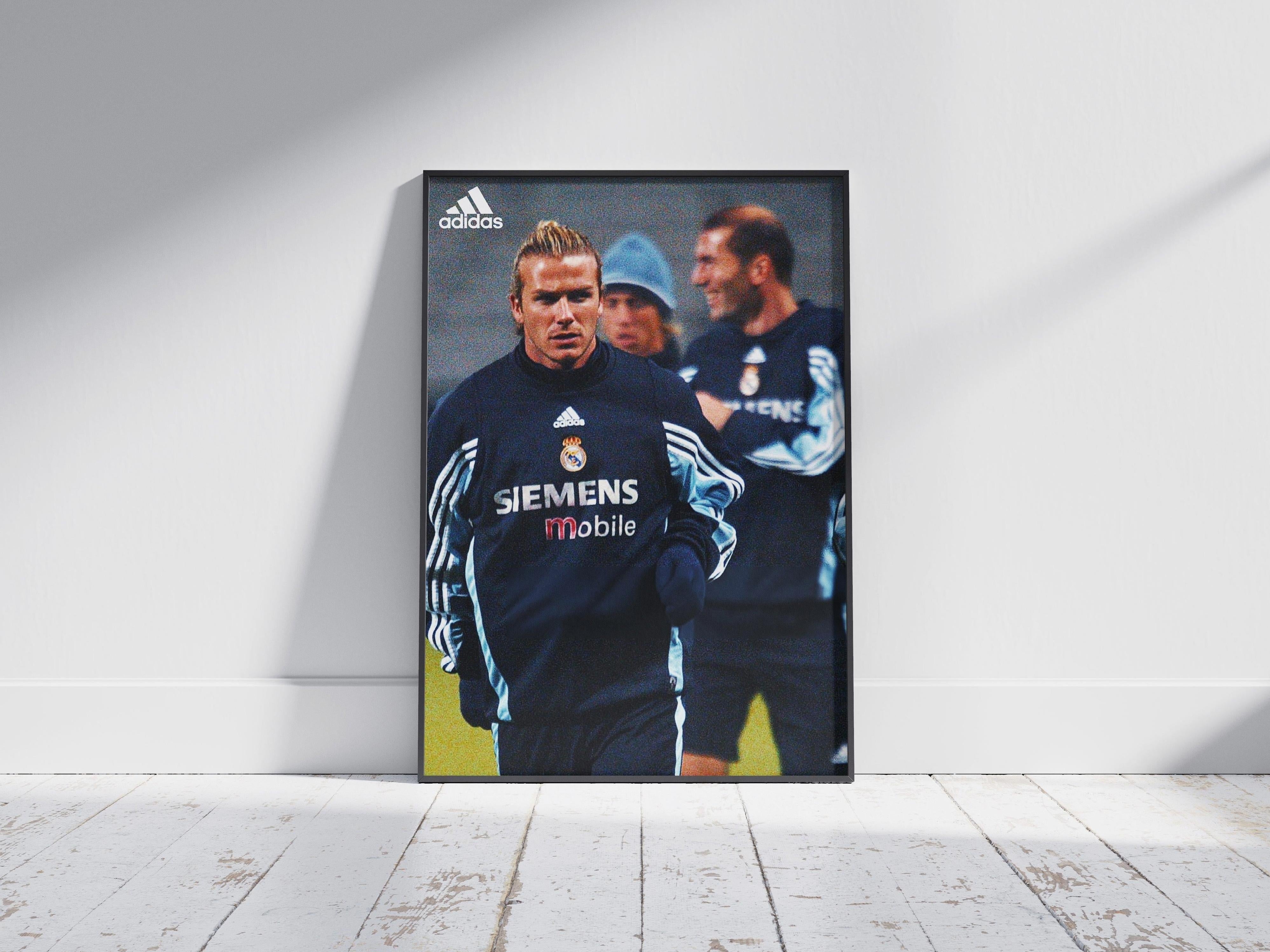 The Beckham x Zidane Poster by Retro1999 features soccer legends in dark blue Real Madrid SIEMENS Mobile training jackets, propped against a white wall on a sunlit wooden floor—a true piece of football history.