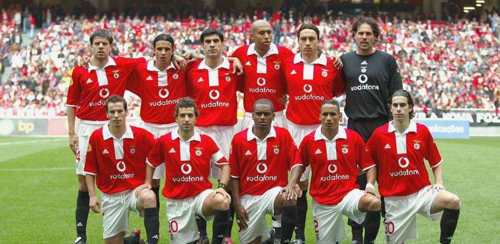 A soccer team in red and white Retro1999 Benfica 2004-05 Home Shirts poses for a group photo on the field before a match, with fans visible in the stadium stands behind them.