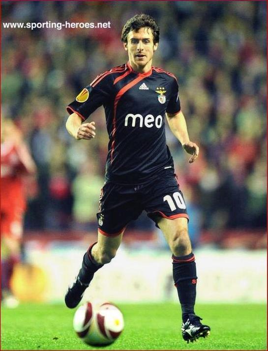 A soccer player wearing a Retro1999 Benfica 2009-10 Away Shirt with number 10 runs on the field during a Primeira Liga match, pursuing the ball, with blurred spectators and players in the background.