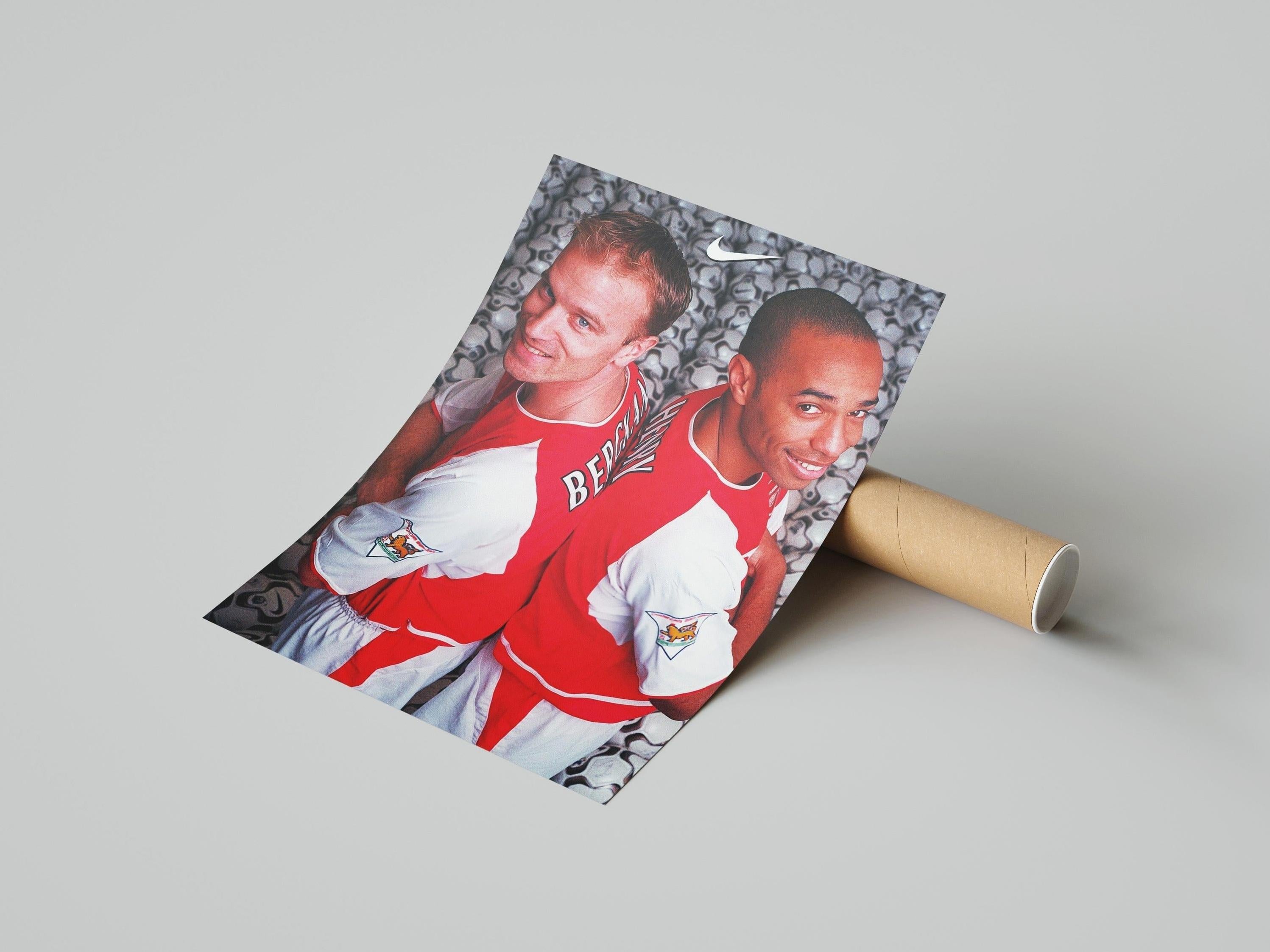 The Retro1999 Bergkamp x Henry Poster shows two male soccer players in red and white jerseys standing back-to-back, smiling, with a background of soccer balls—ideal for football memorabilia or sports decor.
