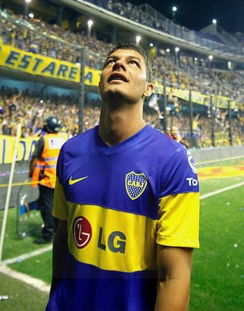 A soccer player wears the Retro1999 Boca Juniors 2010-11 Home Shirt, standing on the field and looking up, with a lively crowd and security staff in the packed stadium behind him.
