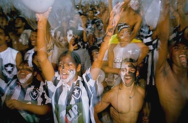 Excited soccer fans, faces painted, cheer and celebrate in Retro1999’s Botafogo 1999-00 Home Shirt, raising their arms and white balloons to support the legendary Brazilian club.