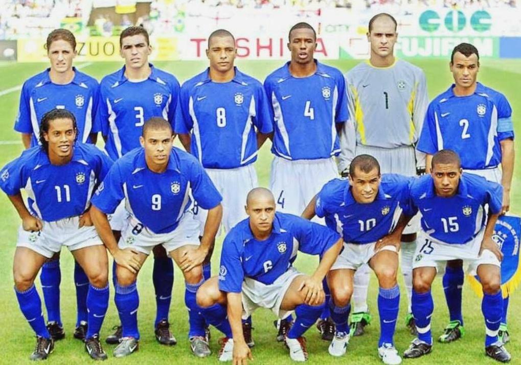 Eleven male soccer players wearing the Retro1999 Brazil 2002 Away Shirt pose for a team photo on the pitch before a match, with stadium fans and advertising boards highlighting this iconic World Cup retro kit.