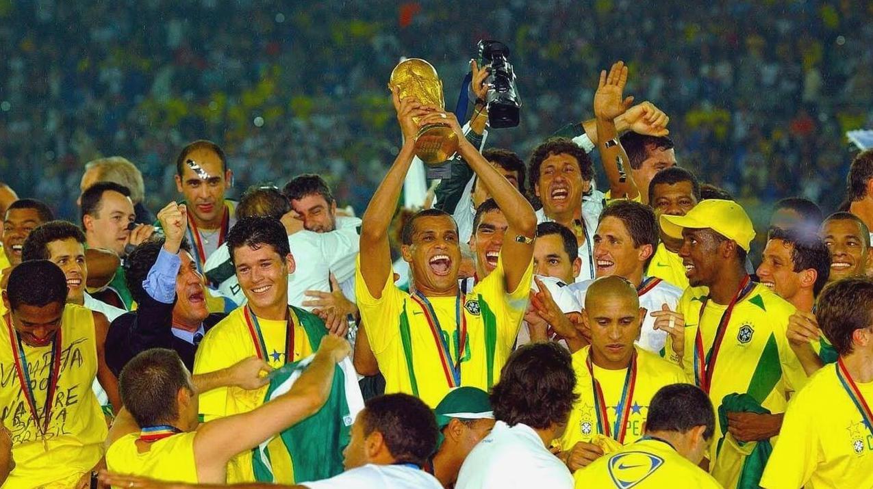 Brazilian soccer players celebrate on the field in the Retro1999 Brazil 2002 Home Shirt, one lifting the FIFA World Cup trophy as teammates cheer with medals around their necks—a legendary football moment.