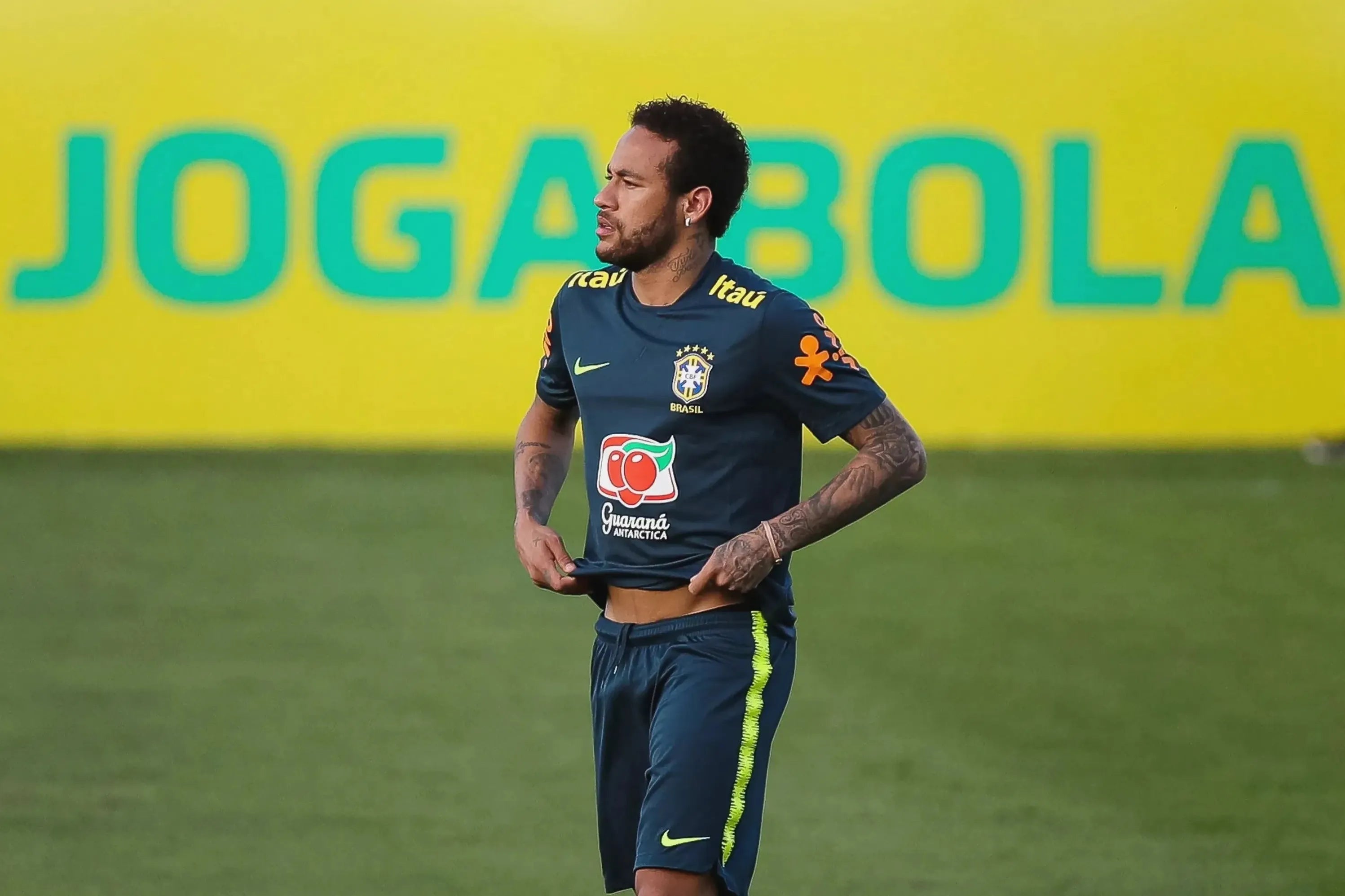 A Brazilian national team player in a Retro1999 Brazil 2019 Training Shirt stands on the field, adjusting his shirt. Behind him, a bright yellow wall features JOGA BOLA in bold green letters.
