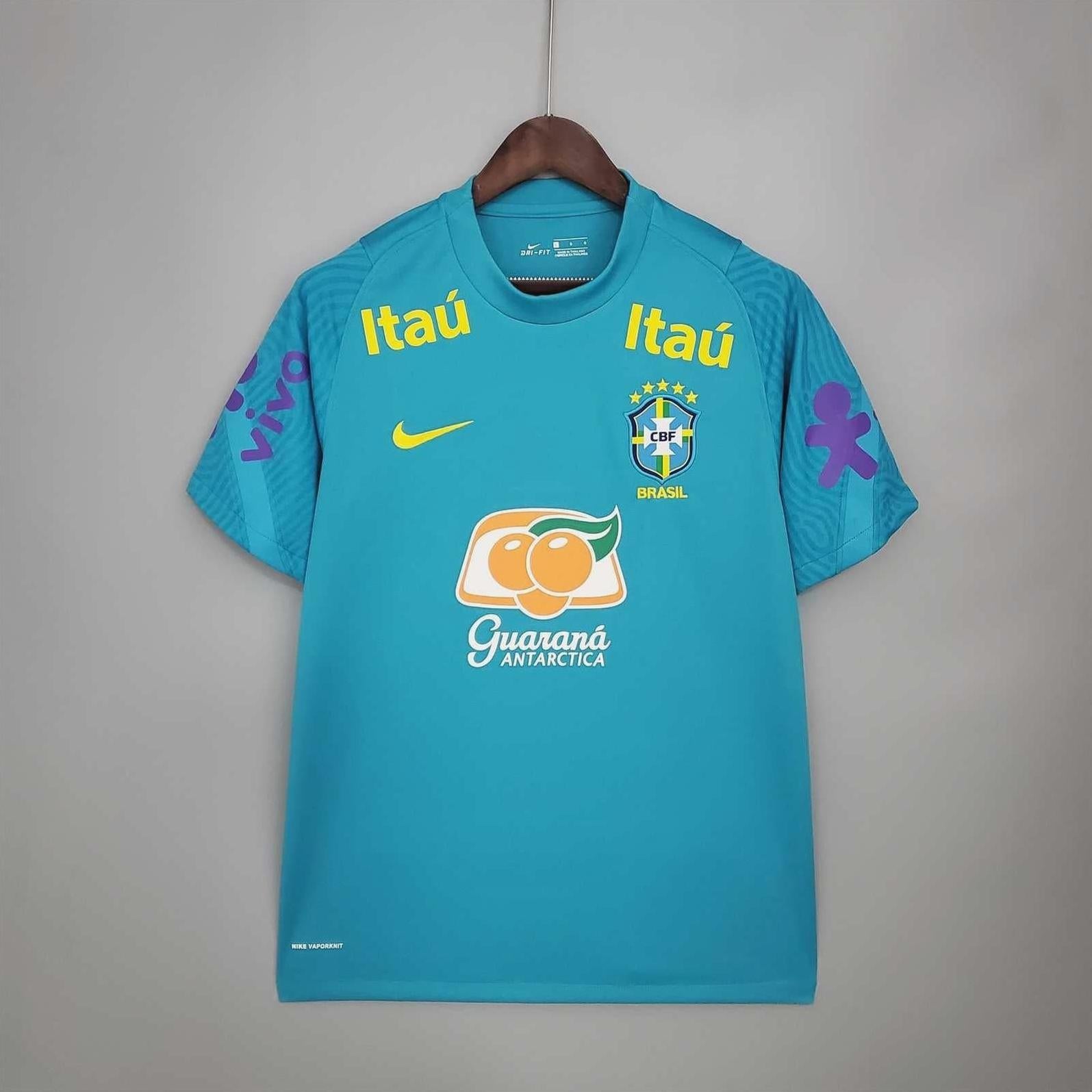 The Brazil 2021 Training Shirt by Retro1999, in teal with sponsor logos and a yellow swoosh, hangs on a wooden hanger. Made from premium fabric, it features the iconic Brazil team crest on the chest for an authentic fan look.
