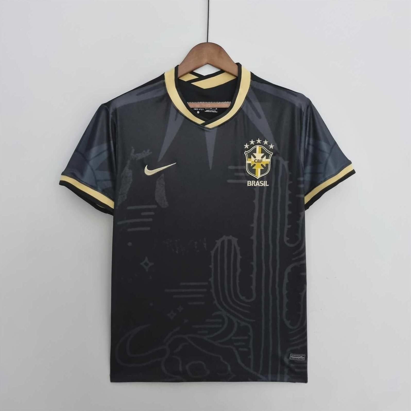 The Retro1999 Brazil 2022 Black Shirt features gold accents, a gold Nike logo, and the Brazil crest with five stars. It hangs on a wooden hanger against a white background, with subtle cactus designs for a retro vibe.