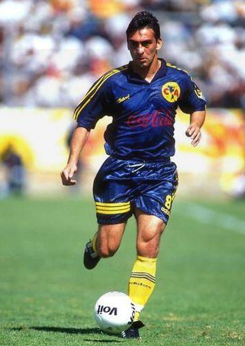 A soccer player in a blue and yellow Retro1999 CF America 1998-99 Away Shirt runs on the field, focused on controlling a white soccer ball during a daytime match.