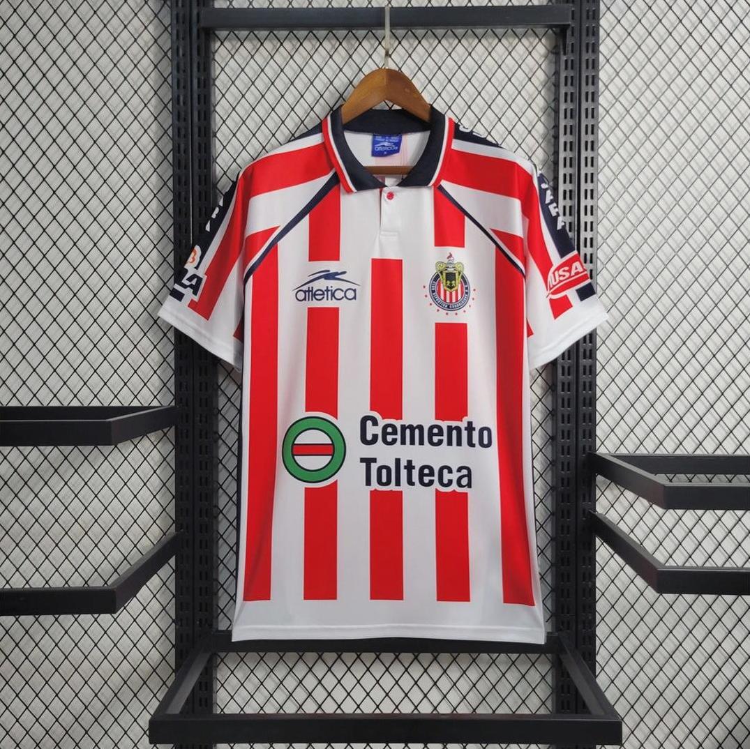 A red and white striped Chivas 2002-03 Home Shirt by Retro1999 with blue accents, a Chivas crest, and Cemento Tolteca sponsor hangs on a hanger against a metal grid wall.