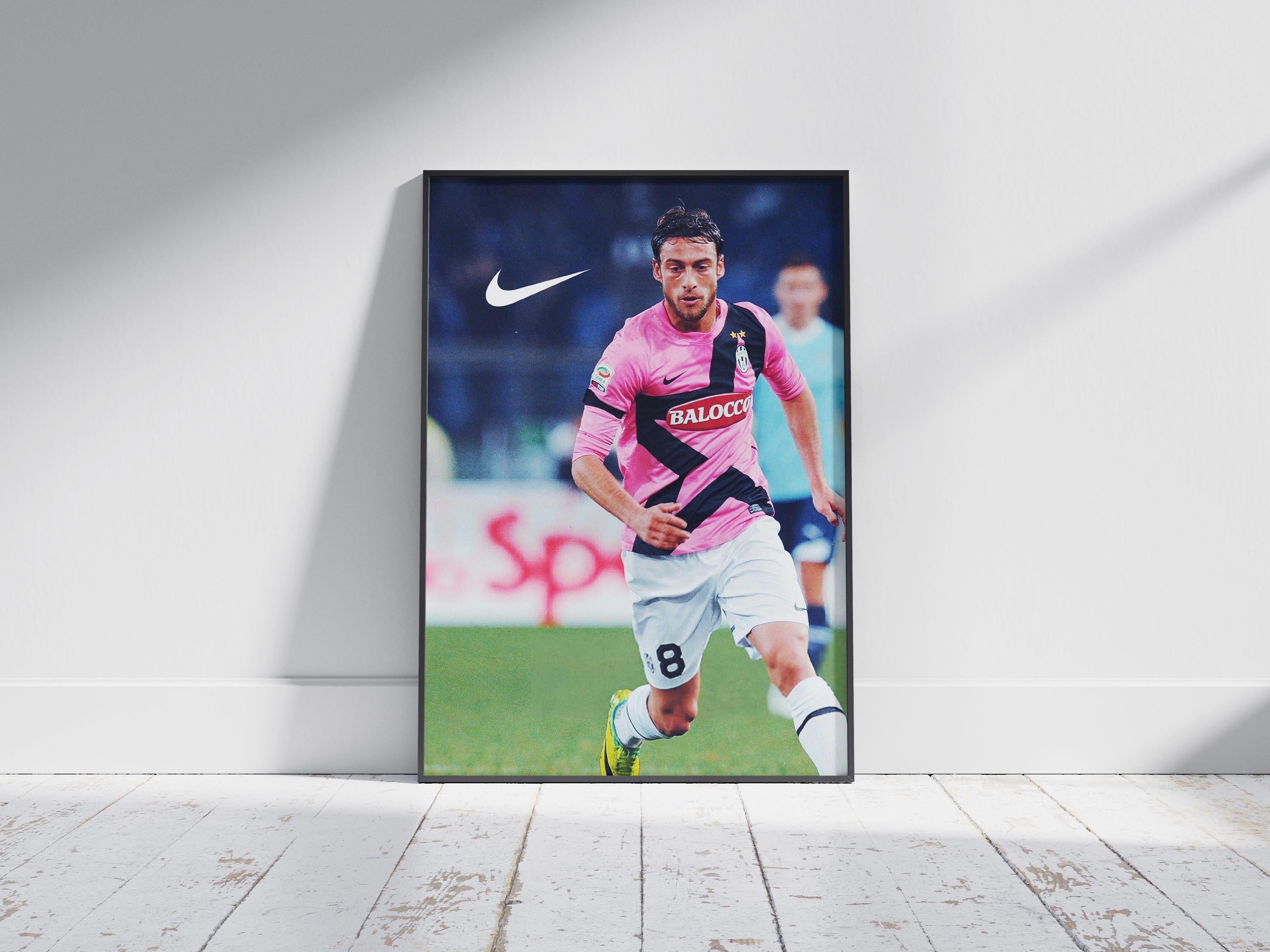 The Retro1999 Claudio Marchisio x Juventus 2011/12 Poster shows Marchisio in a pink and black kit running on the field, set against a white wall and light wooden floor—perfect Serie A memorabilia for fans.