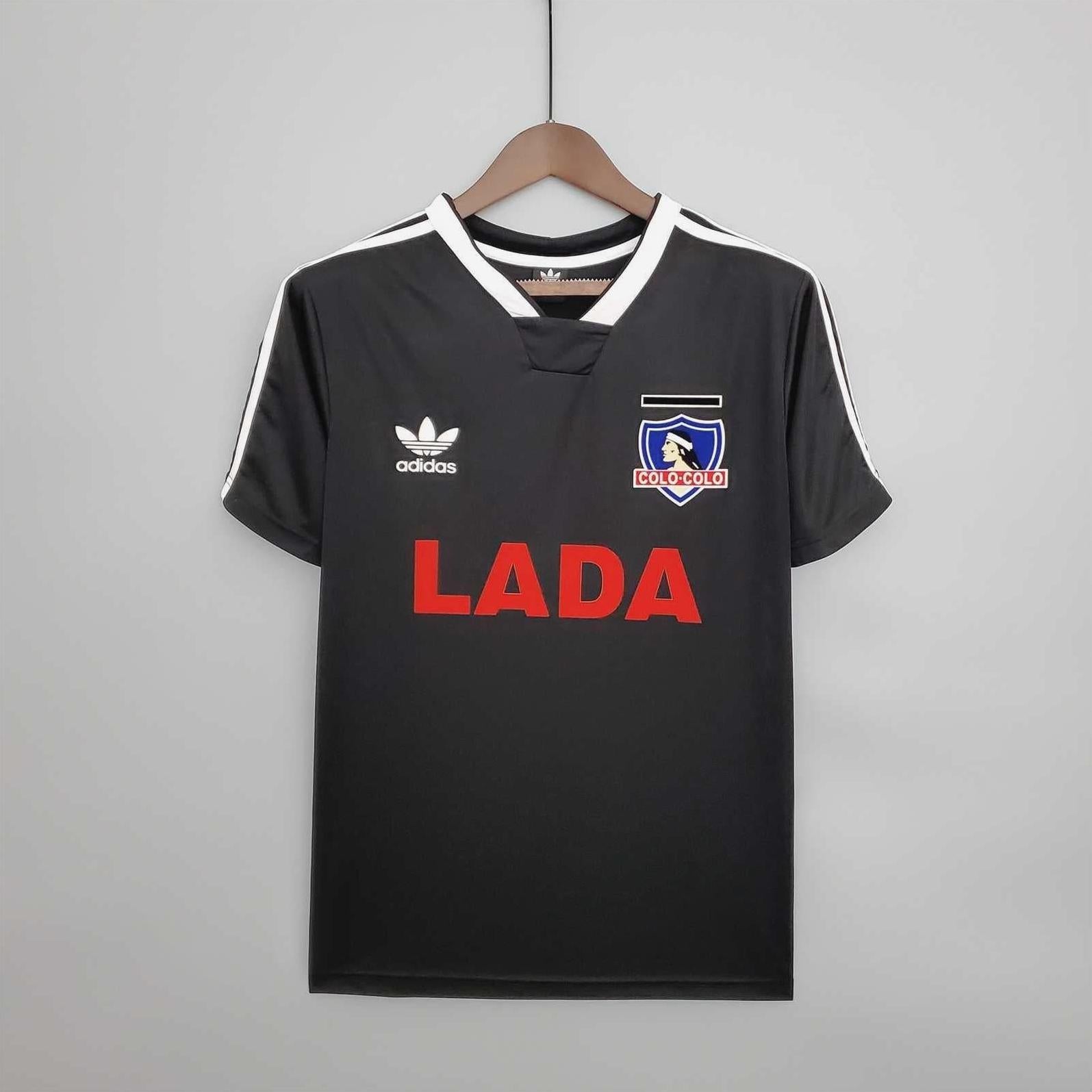 A Colo Colo 1991 Away Shirt by Retro1999 in black with white accents, featuring the Adidas logo, club crest, and bold red LADA lettering on the front, displayed on a hanger against a plain light background.