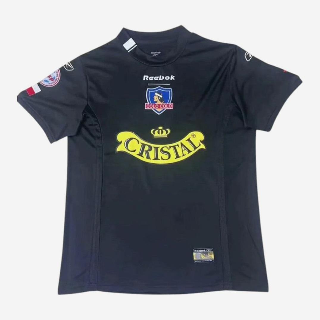 A black Colo Colo 2004 Away Shirt by Retro1999 with yellow Cristal sponsor, Reebok branding, team and league sleeve patches. This retro kit shows an authentic design and is laid flat on a white background.