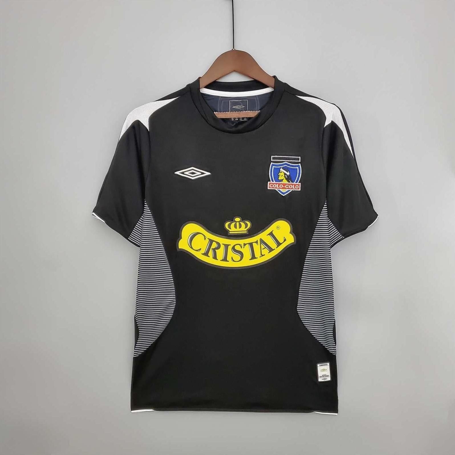 The Retro1999 Colo Colo 2006 Away Shirt in black with white and gray accents features the Umbro logo, club crest, and bold yellow CRISTAL sponsor. This premium retro kit is shown on a hanger against a gray background.