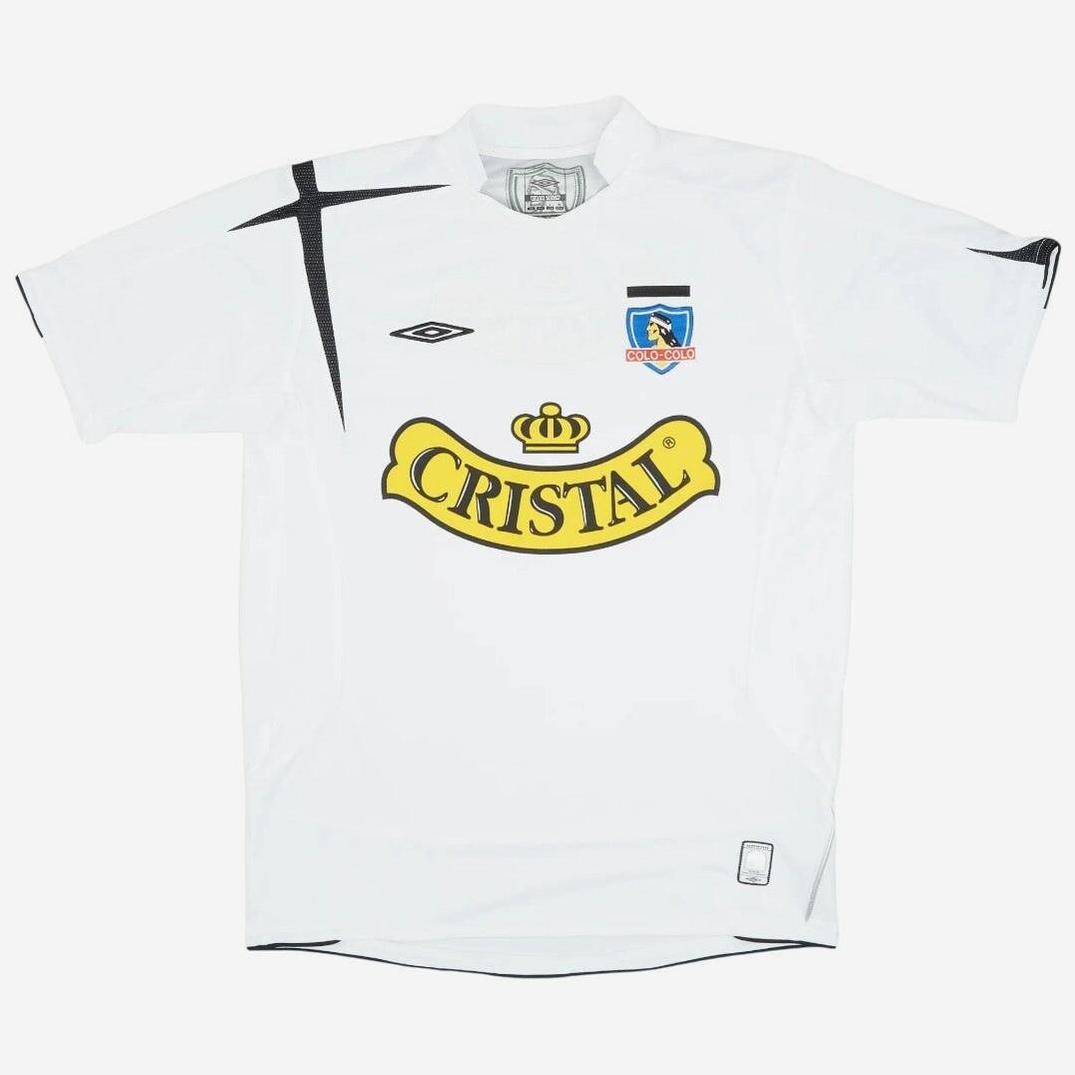 The Colo Colo 2006 Home Shirt by Retro1999 is a white soccer jersey with black accents, the Umbro logo, club crest on the chest, and a large yellow Cristal sponsor—an iconic piece in football history.