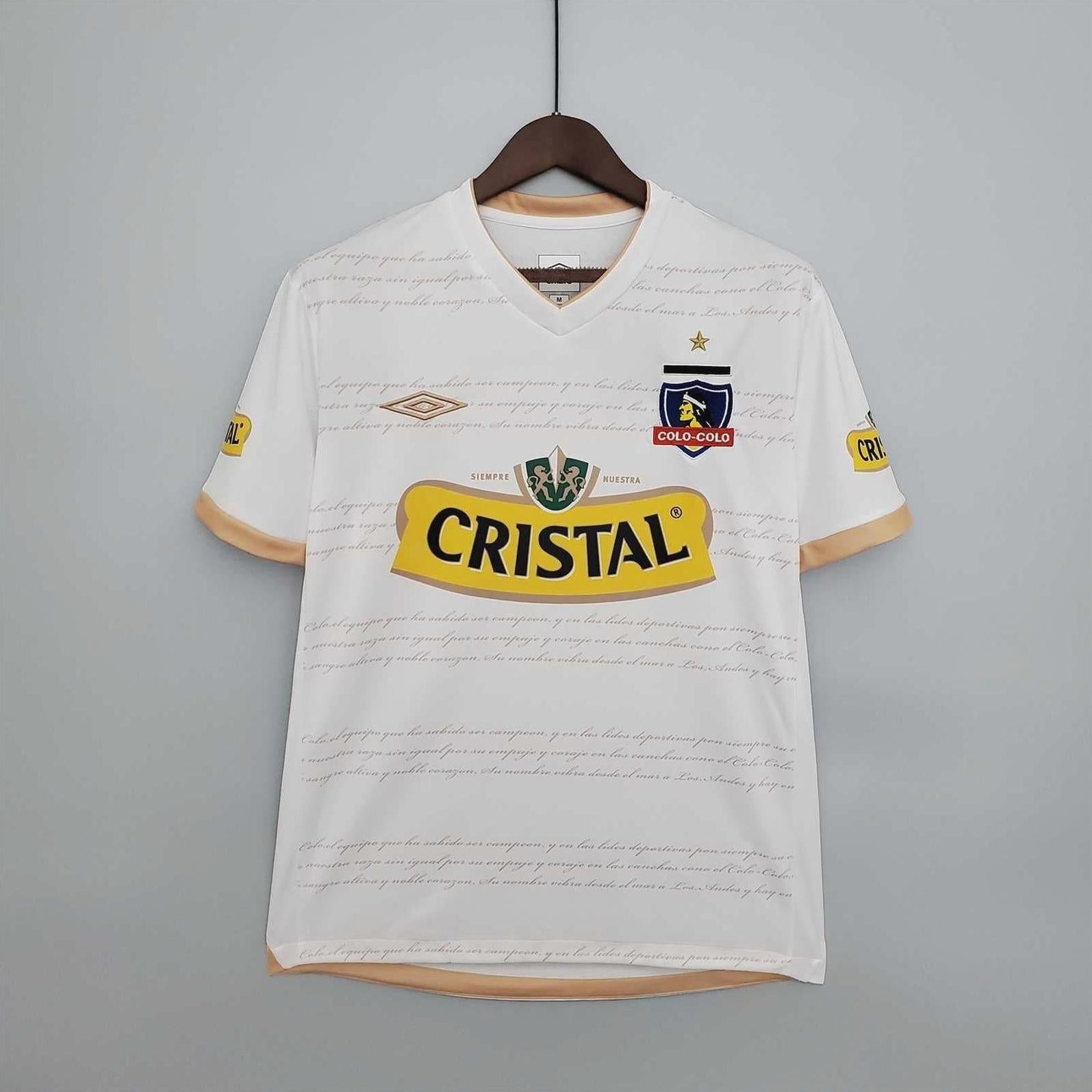 The Retro1999 Colo Colo 2011 Home Shirt is white with beige accents, featuring the club crest and branding. This retro kit shows  team colors and faint script text on the front, displayed on a hanger.