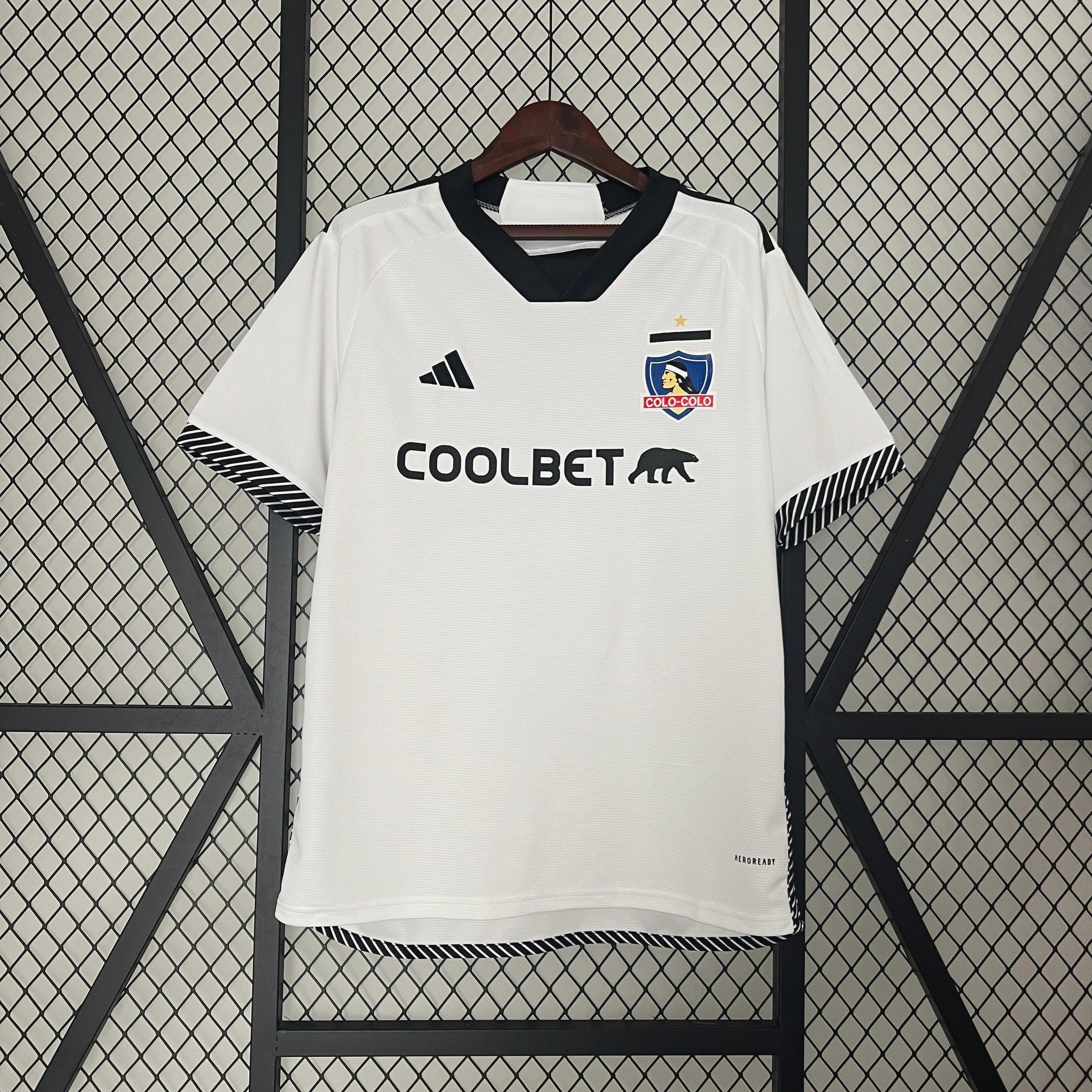 The Colo Colo 2024-25 Home Shirt by Retro1999, in white with black collar and cuffs, hangs on a hanger. The  design displays the club crest, Adidas logo, and COOLBET sponsor bear graphics against a wire fence backdrop.