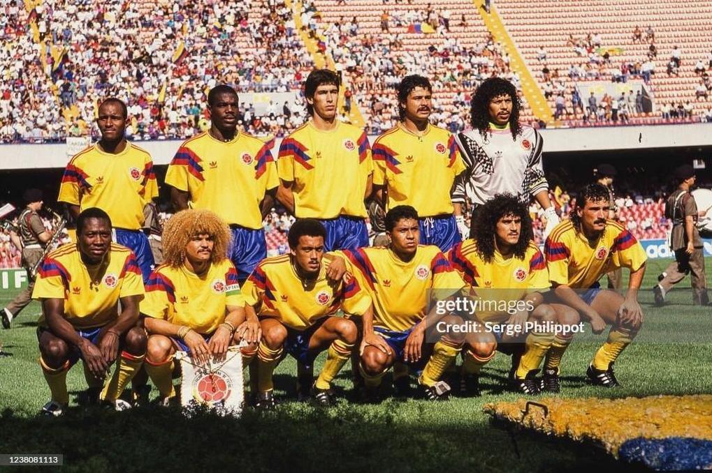 A men’s soccer team poses in Retro1999’s Colombia 1990 Home Shirt—yellow, blue, and red retro kit—before a match in a crowded stadium, evoking memories of the iconic design as players stand and crouch for a group photo.