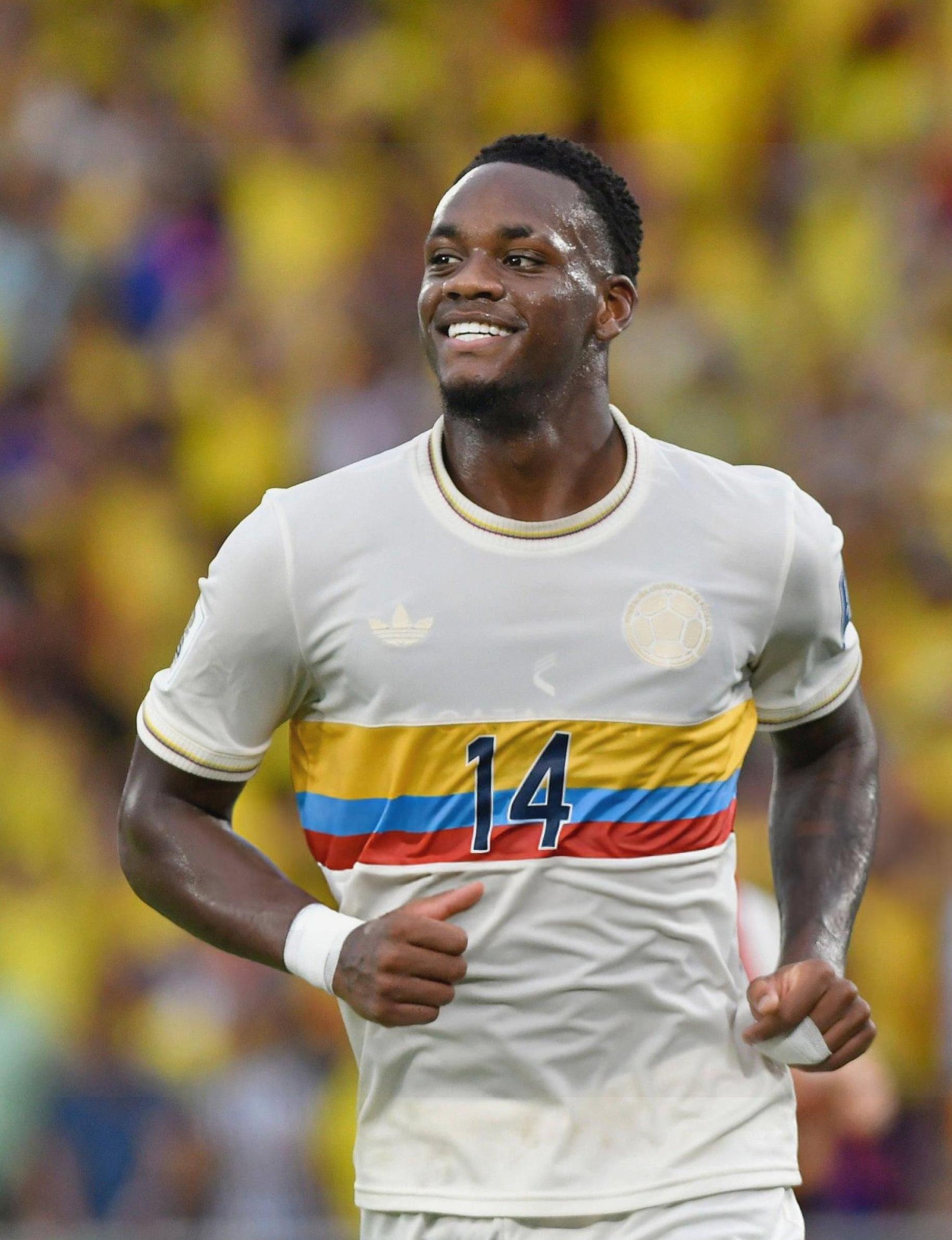 Wearing the Retro1999 Colombia 2024 Centenary Kit—a white jersey with yellow, blue, and red stripes and number 14—a soccer player smiles while running on the field, with blurred yellow-clad fans celebrating Colombian football.