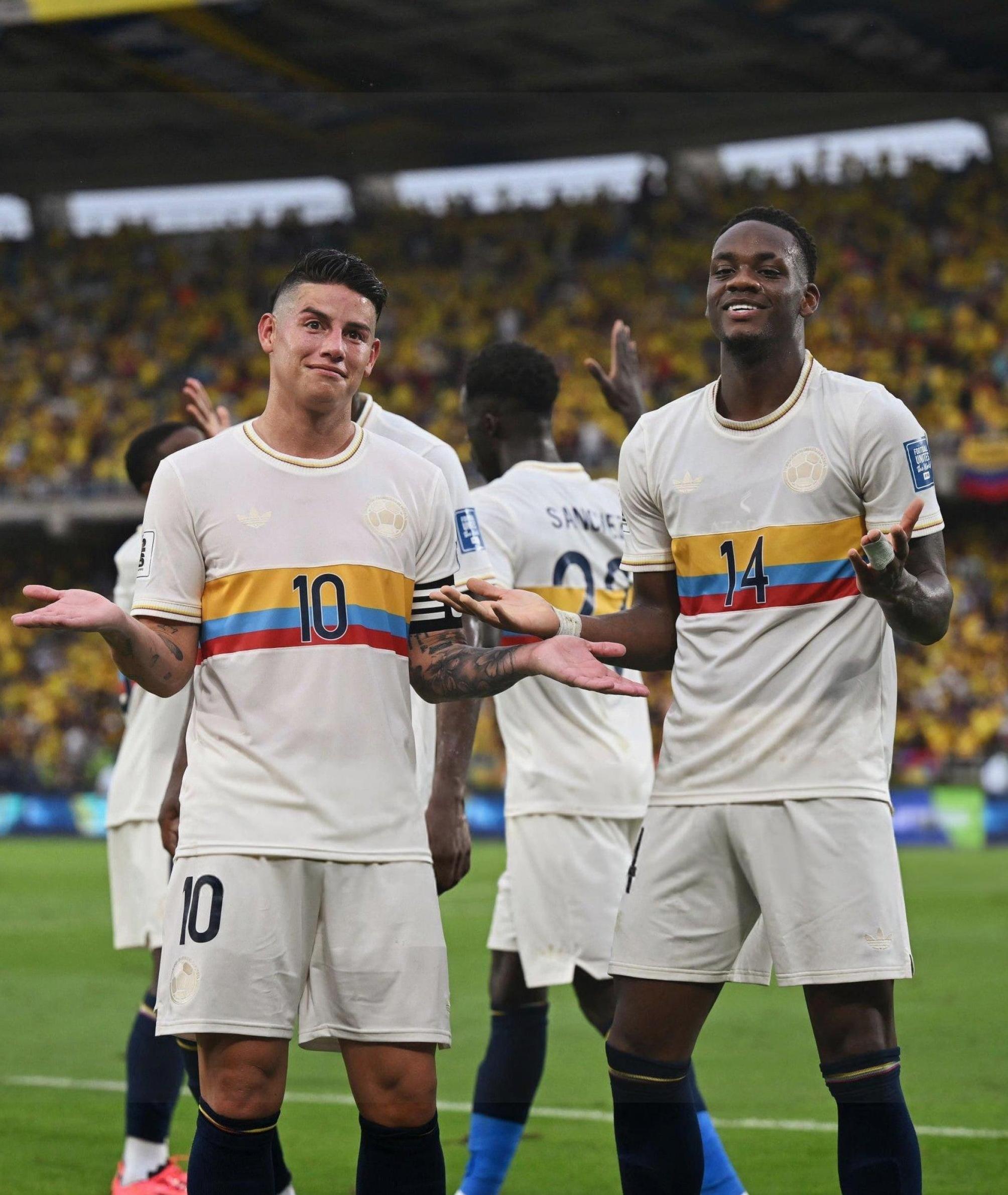 Two male players in Retro1999’s Colombia 2024 Centenary Kit, white with colorful stripes and numbers 10 and 14, stand on the field smiling and shrugging, as a stadium crowd in yellow celebrates Colombian football excellence.