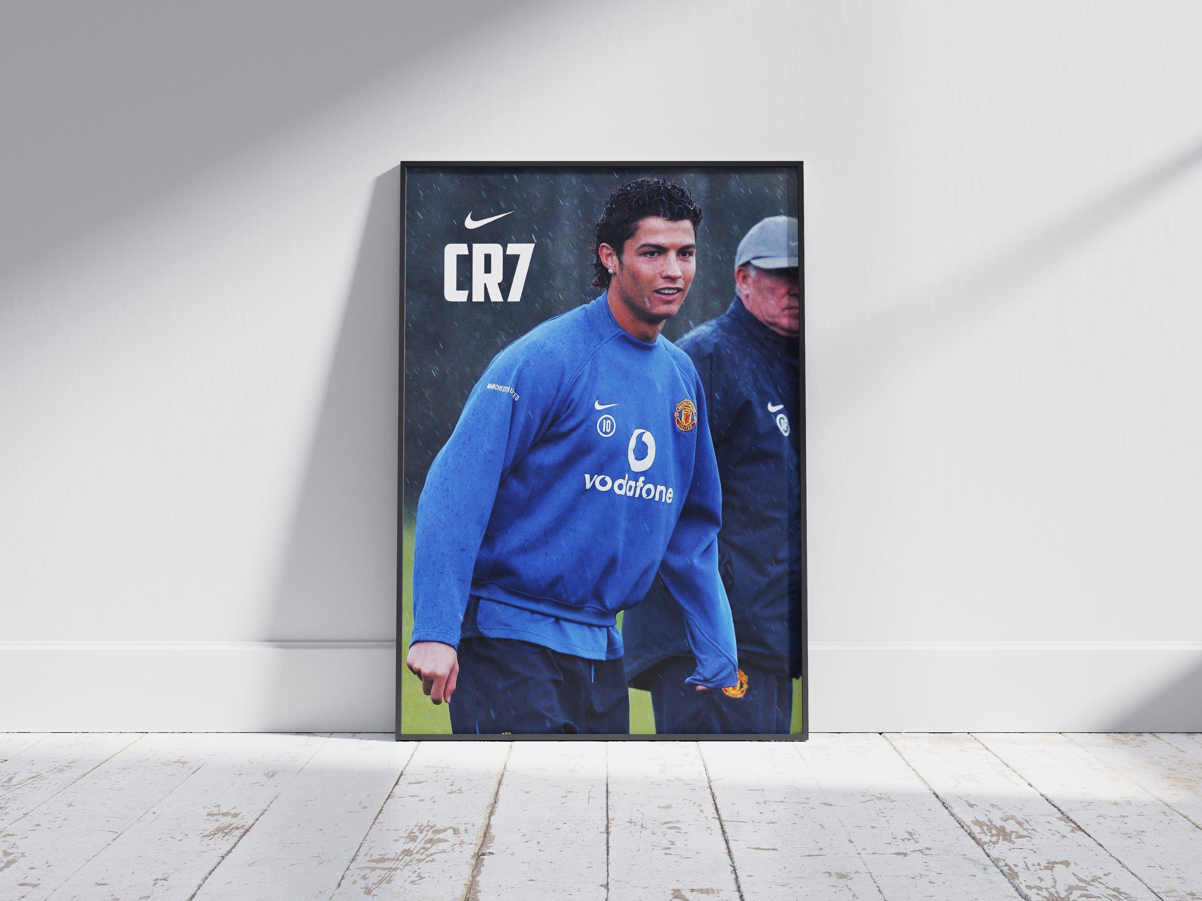 The Retro1999 CR7 x Sir Alex Poster features a male soccer player in a blue Manchester United training top with CR7 and Nike logos, leaning against a white wall on a wooden floor—a stylish tribute to Manchester United history.