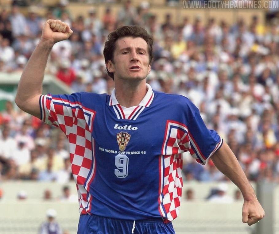 Wearing the Retro1999 Croatia 1998 Away Shirt, the player celebrates with a raised fist on the field as a blurred crowd roars—a vibrant tribute to classic Croatian football glory in this iconic blue and red checkered kit.