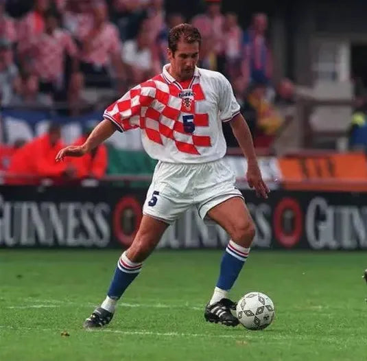 A soccer player in the Retro1999 Croatia 1998 Home Shirt, with white shorts and blue socks, controls the ball during a World Cup 1998 match. The number 5 retro kit catches attention as fans watch from the stands.
