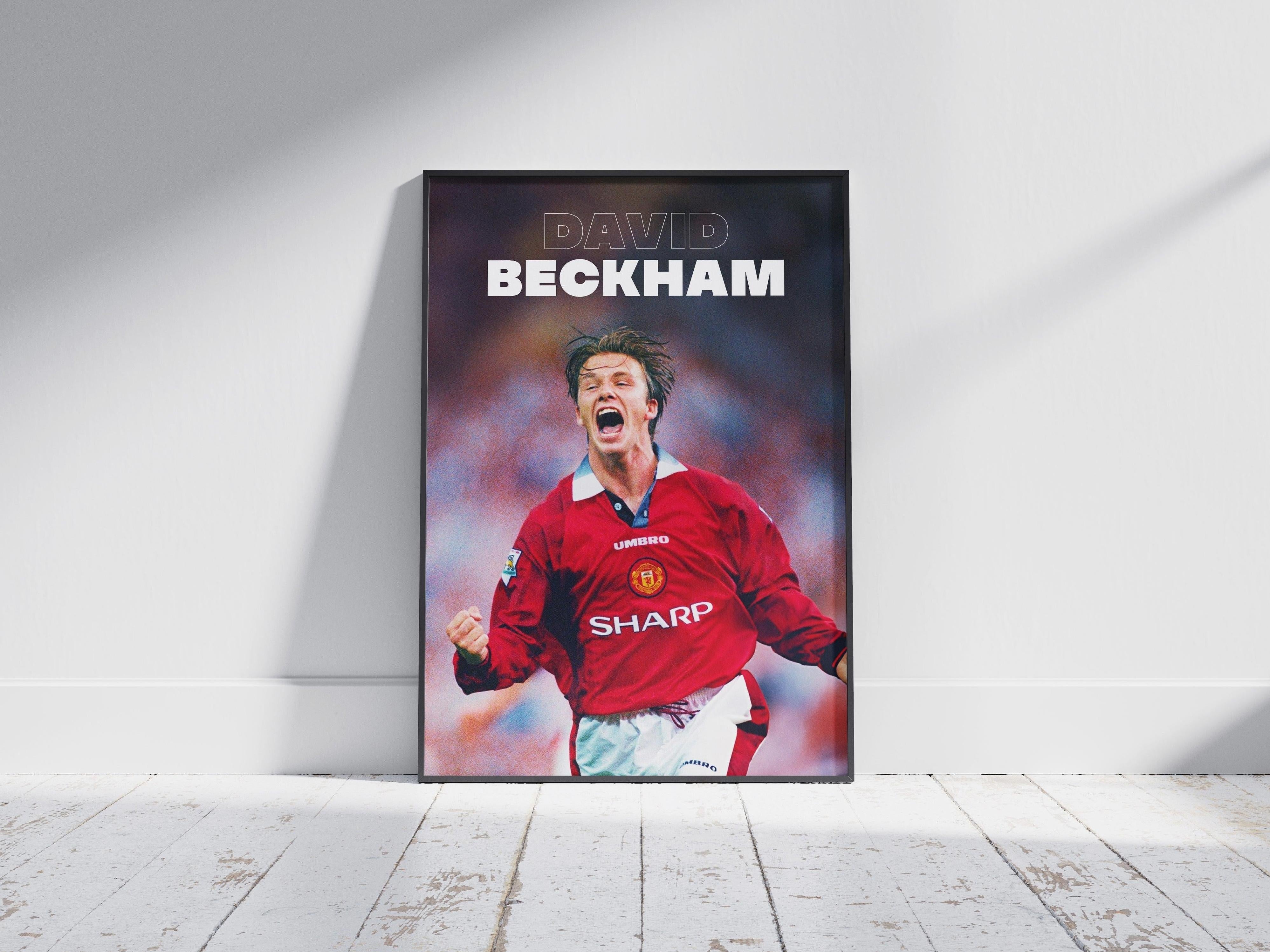 The David Beckham x Manchester United Poster by Retro1999 features the legend in a red jersey celebrating, with his name at the top, framed and displayed on a light wooden floor against a white wall.
