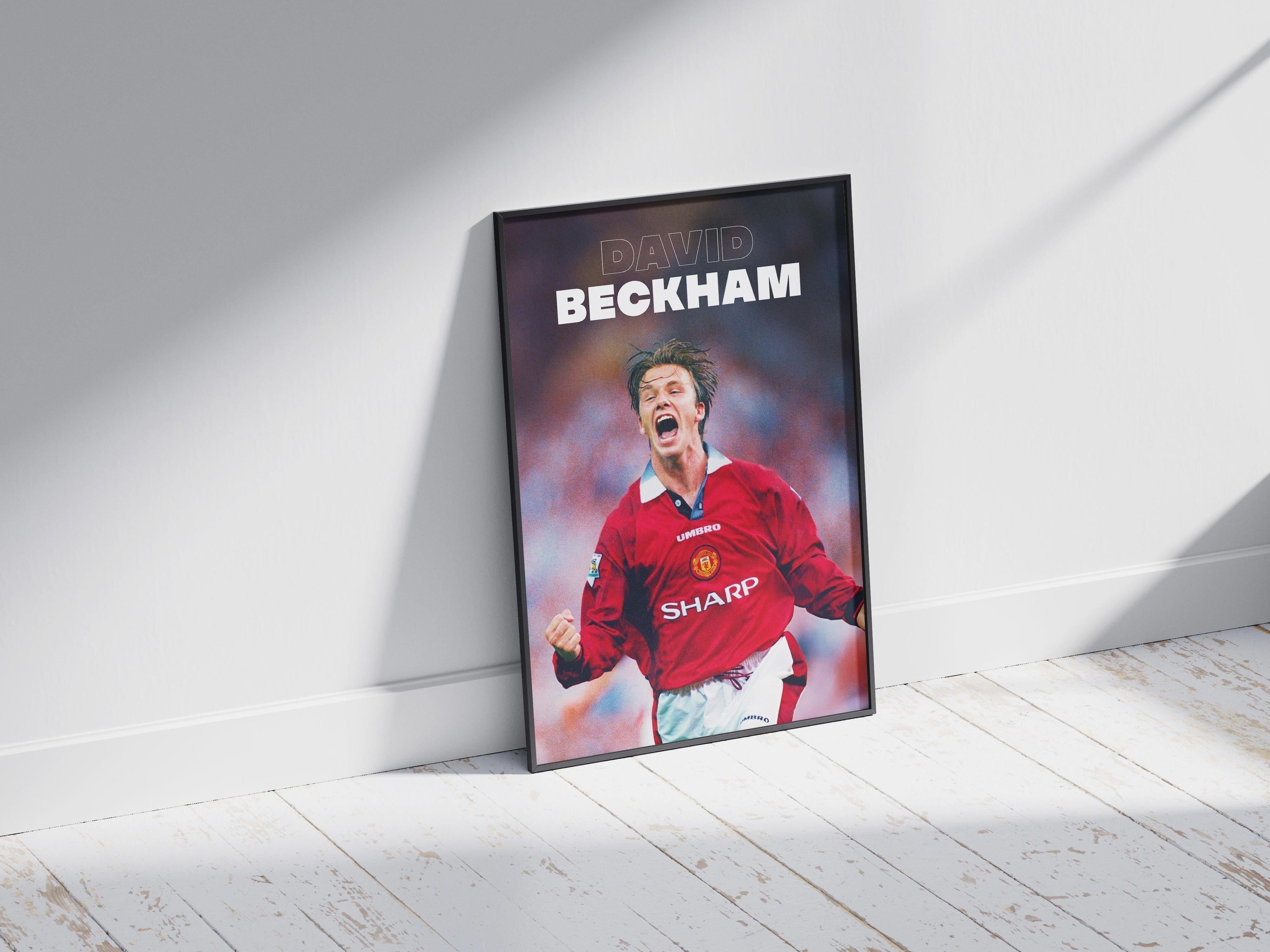 The Retro1999 David Beckham x Manchester United Poster features Beckham in a red jersey, celebrating with clenched fists. DAVID BECKHAM appears at the top. The framed poster leans against a white wall on a wooden floor.