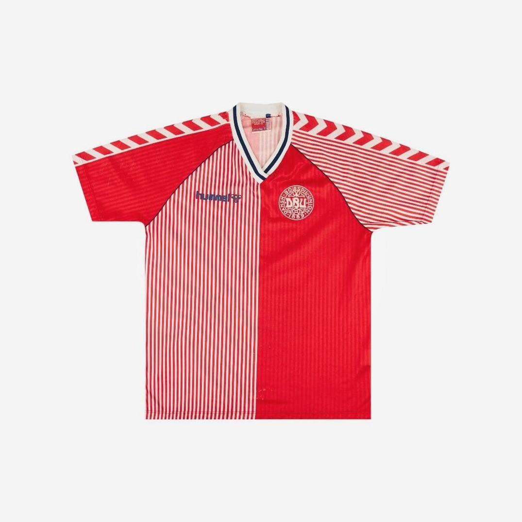 The Denmark 1986 Home Shirt by Retro1999 features bold red and white halves, vertical stripes, chevron shoulder patterns, the team logo, and a blue sponsor on the chest—celebrating classic football history.