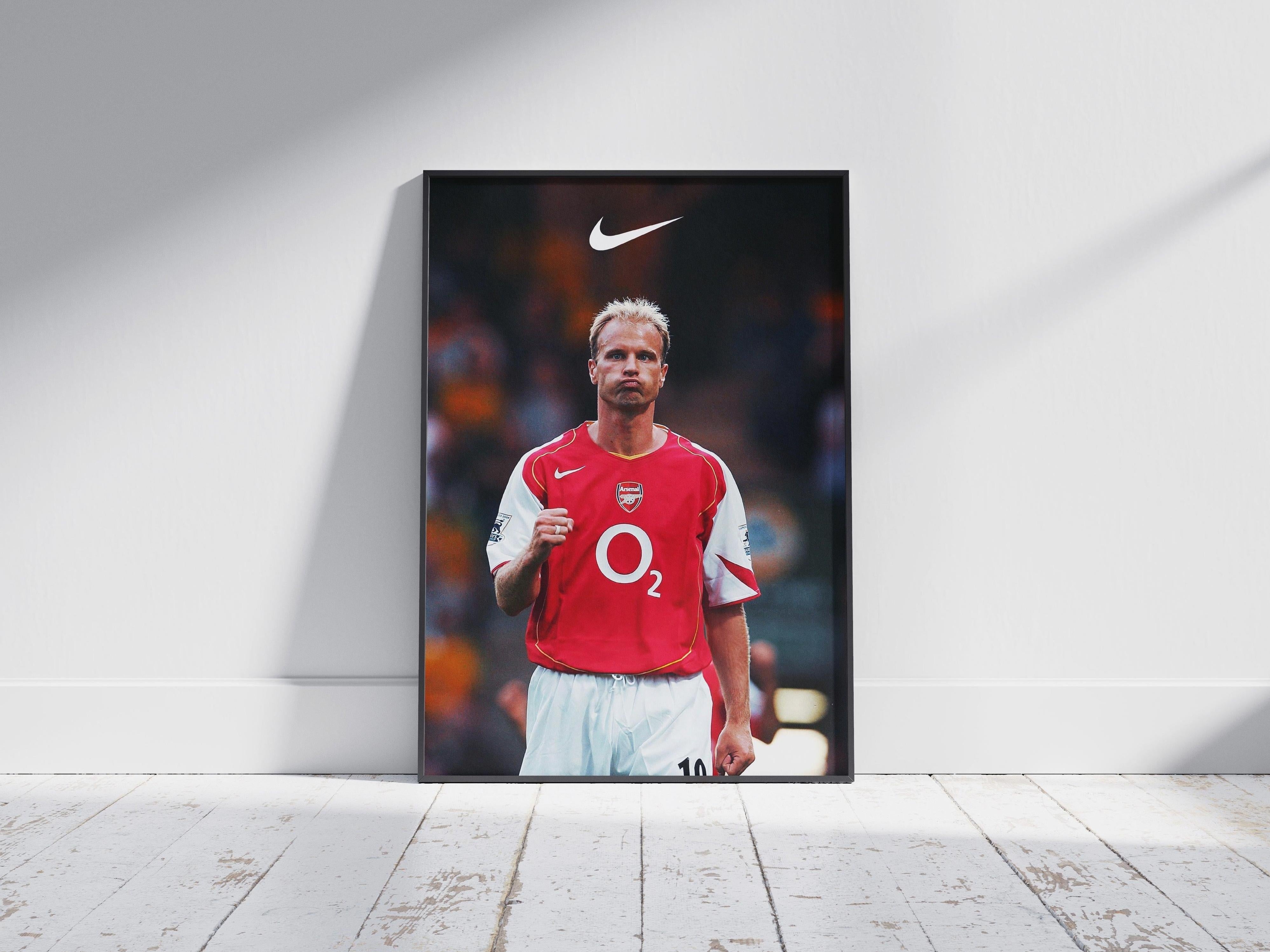 The Retro1999 Dennis Bergkamp x Arsenal Poster features Bergkamp in the classic O2 jersey, framed and set against a white wall with sunlight and a wooden floor—an ideal piece for fans honoring Arsenals football legacy.