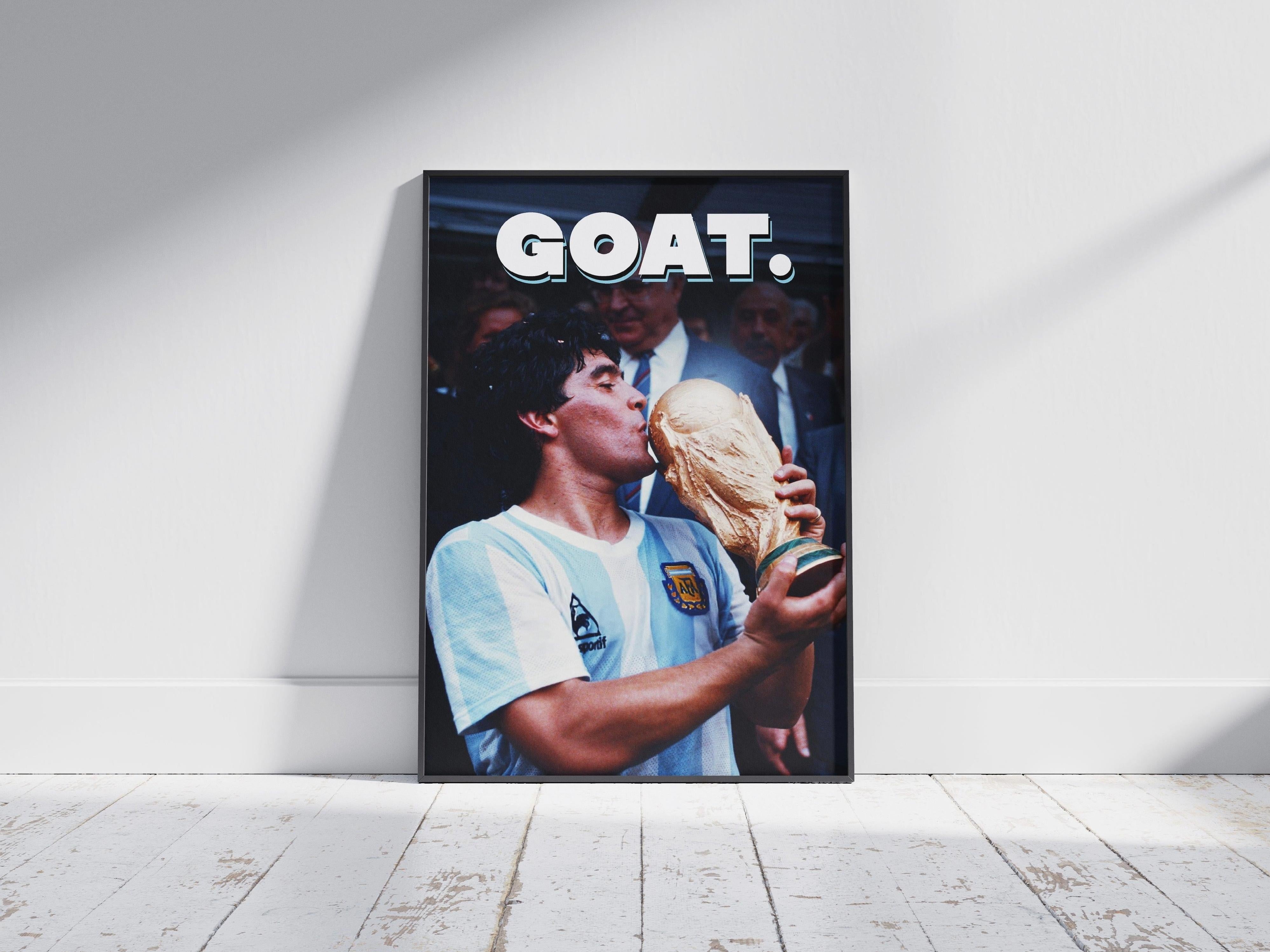 The Diego Maradona GOAT Poster by Retro1999 features Maradona in an Argentina jersey kissing the 1986 FIFA World Cup trophy, with bold white GOAT. text above, framed and displayed on a white wooden floor against a plain wall.