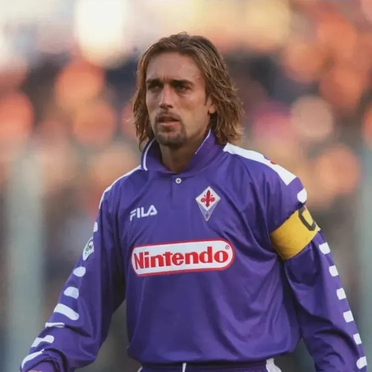 Wearing the Retro1999 Fiorentina 1998-99 Home Shirt with its famous Nintendo sponsor and a yellow captains armband, a soccer player stands on the pitch during a Serie A match, highlighting this iconic retro kit.